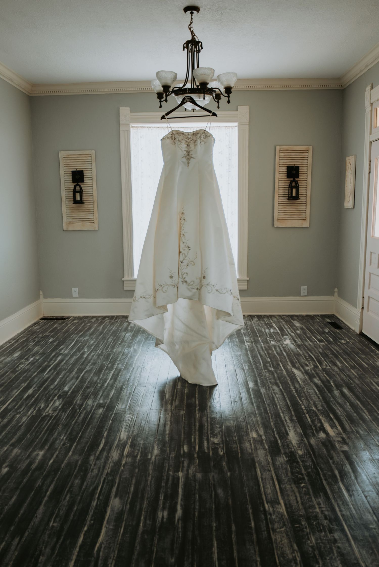 A stunning white wedding dress hangs elegantly from a chandelier in a room with gray walls and dark hardwood floors.