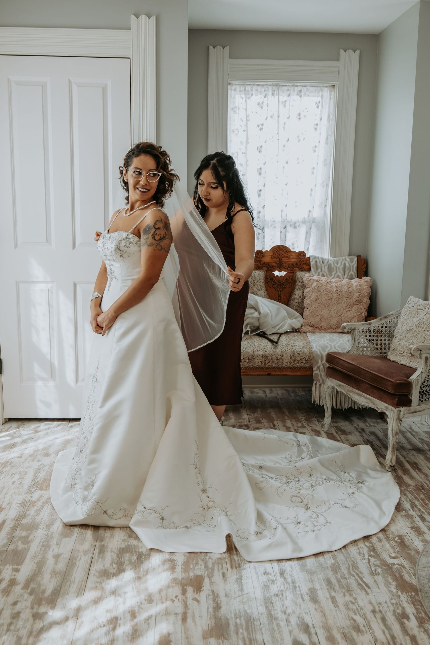 A wedding dress fitting moment in a sunlit room with vintage furniture and lace curtains decorating the intimate space.