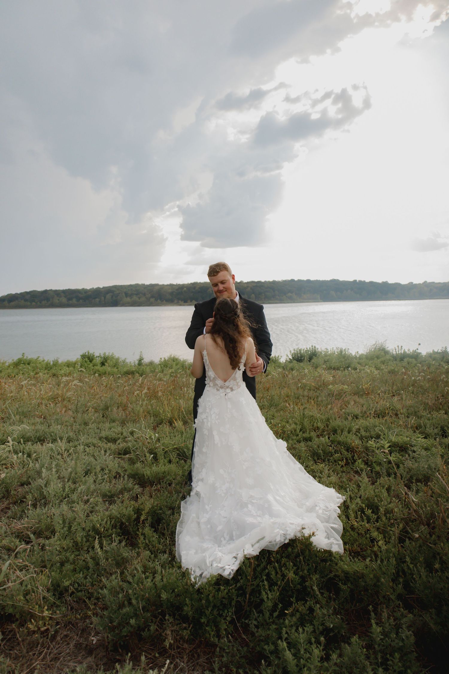 A couple in wedding attire embraces while standing in a grassy field overlooking a serene lake on their special day.
