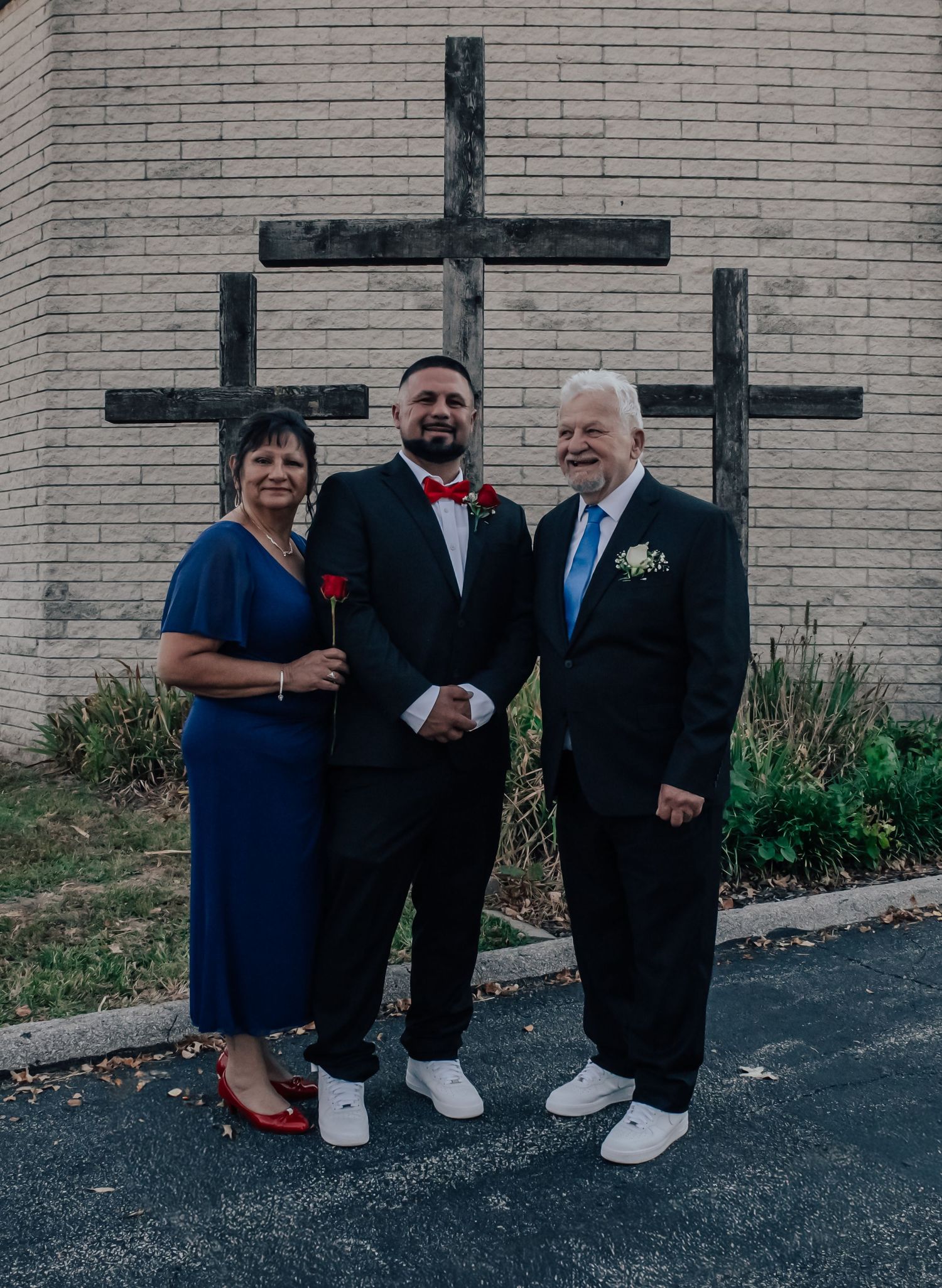 Three people in formal attire stand together in front of three wooden crosses mounted on a light brick church wall.