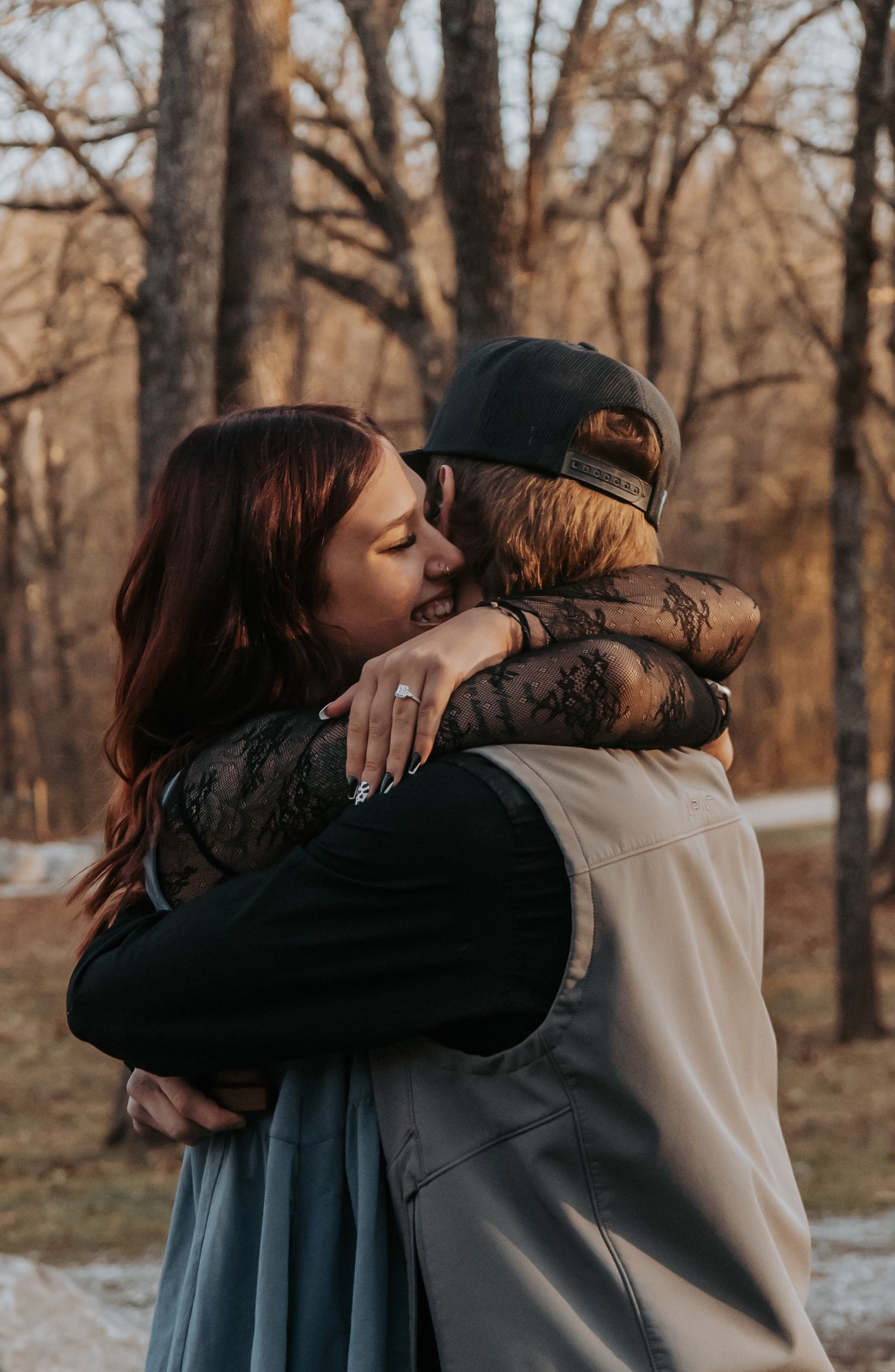 Two people share an emotional embrace in a winter forest setting with bare trees in the background.