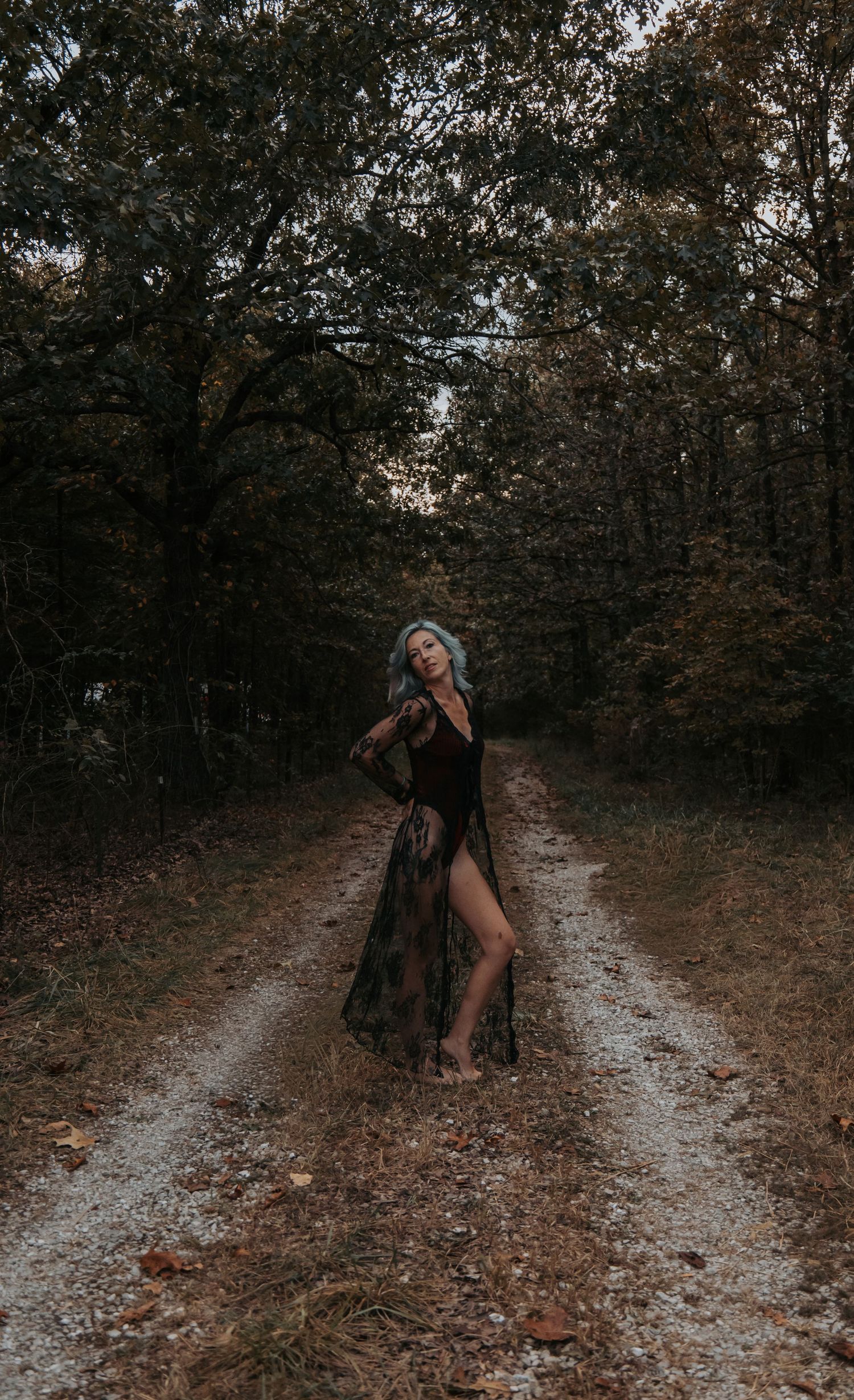 A figure in a dark dress dances gracefully along a forest path covered in fallen autumn leaves.