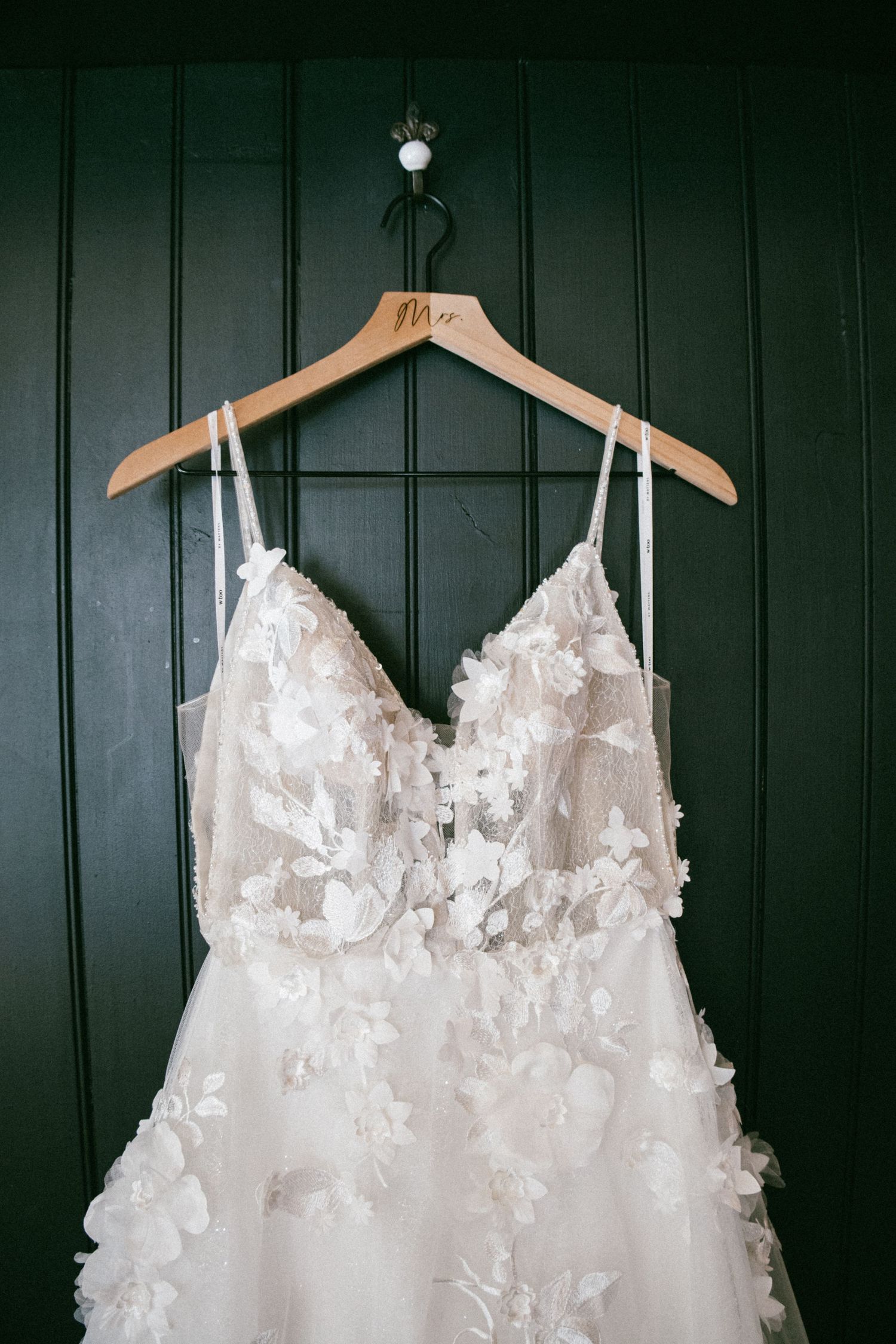 Three elegant white wedding dresses with delicate floral appliques hanging against a dark green wall.