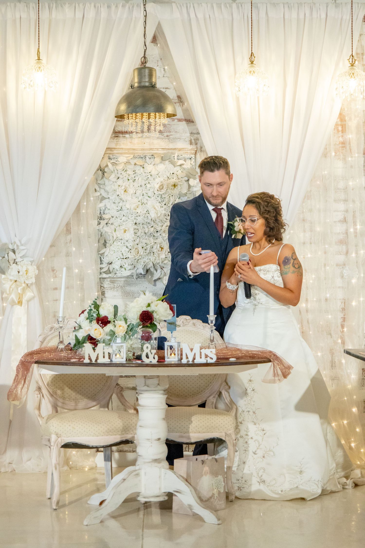 A romantic moment between newlyweds looking at their phones near an elegantly decorated wedding table with sheer curtains.