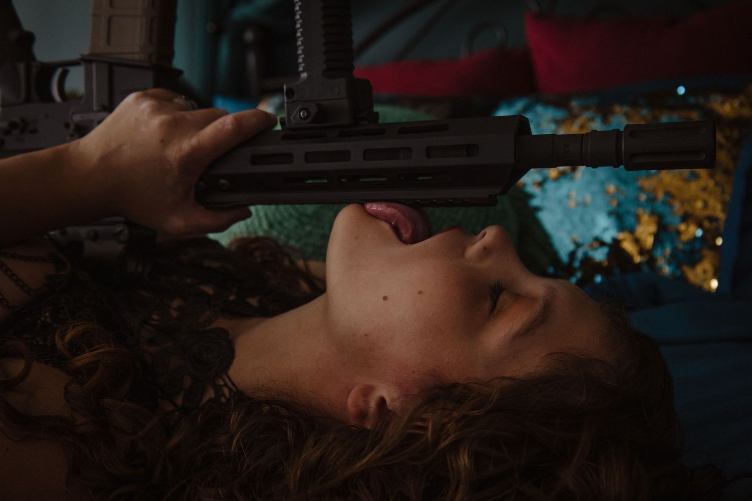 Person lying down with eyes closed while holding a rifle in a dimly lit setting with blue and yellow lights in background.