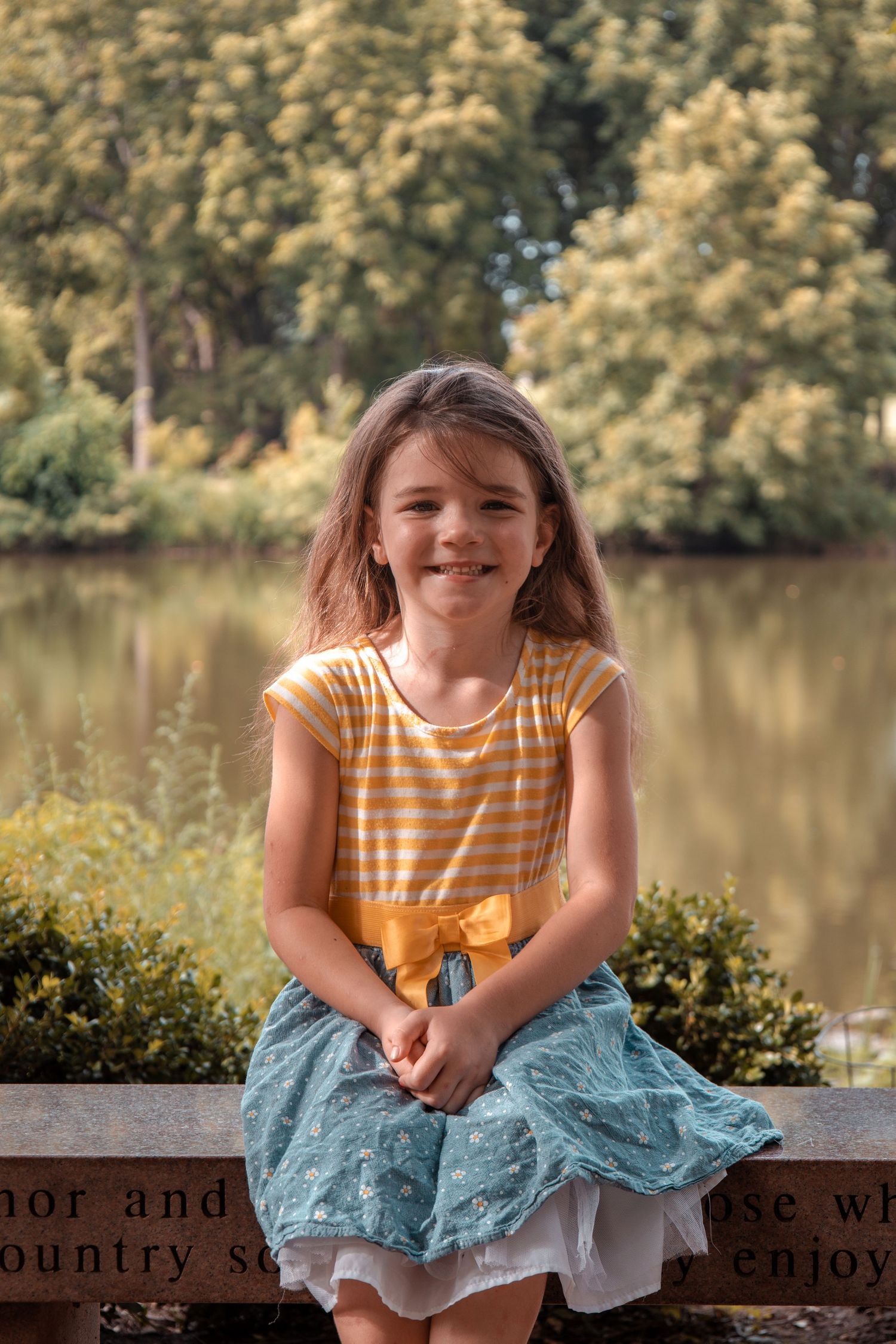 A child in a yellow striped shirt and blue skirt sits on a stone wall by a serene lake surrounded by autumn trees.