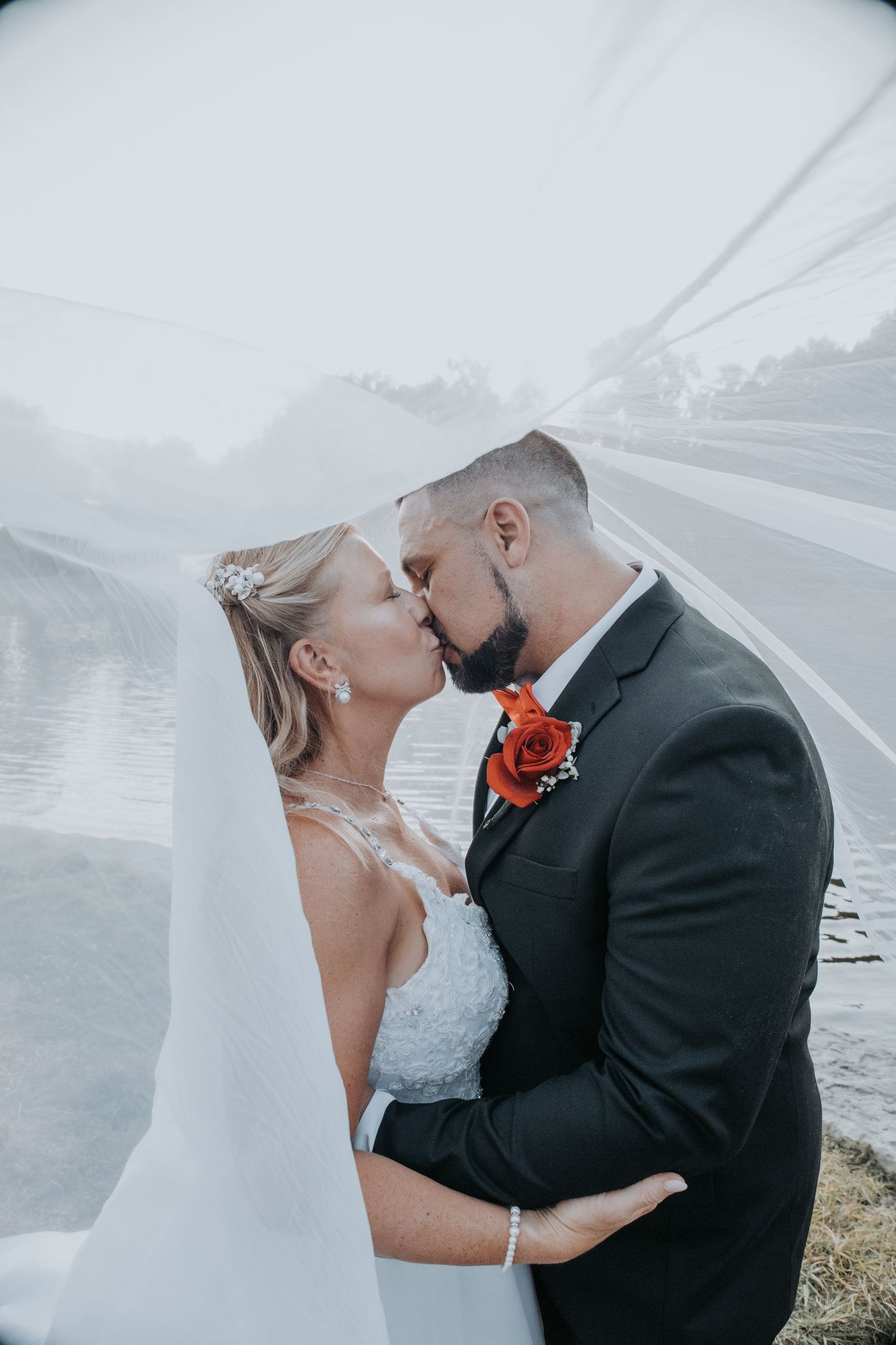 A romantic wedding couple shares a tender kiss under a flowing white veil against a misty mountain backdrop.