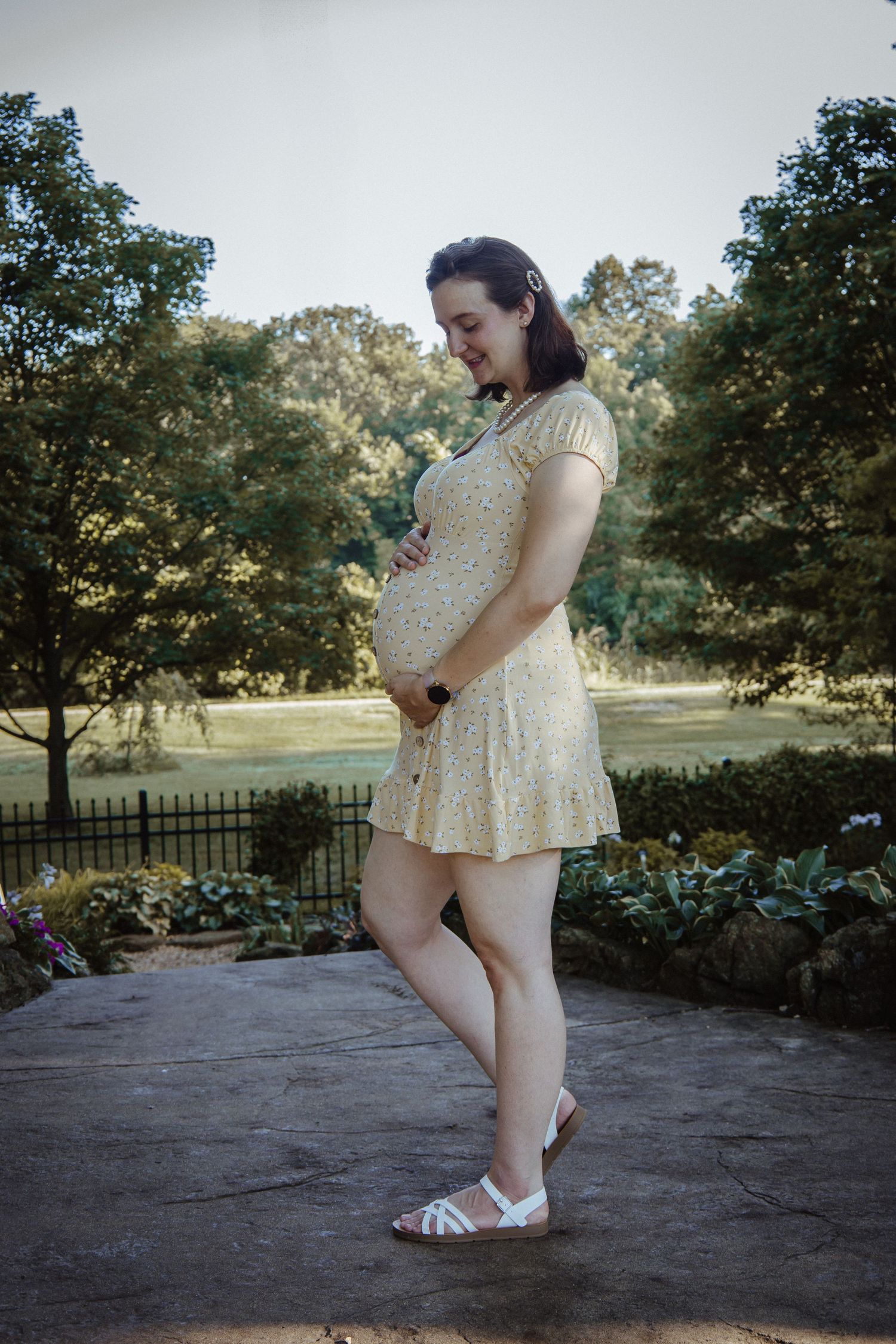 A pregnant woman in a cream lace dress stands on a stone patio in a garden setting during golden hour.