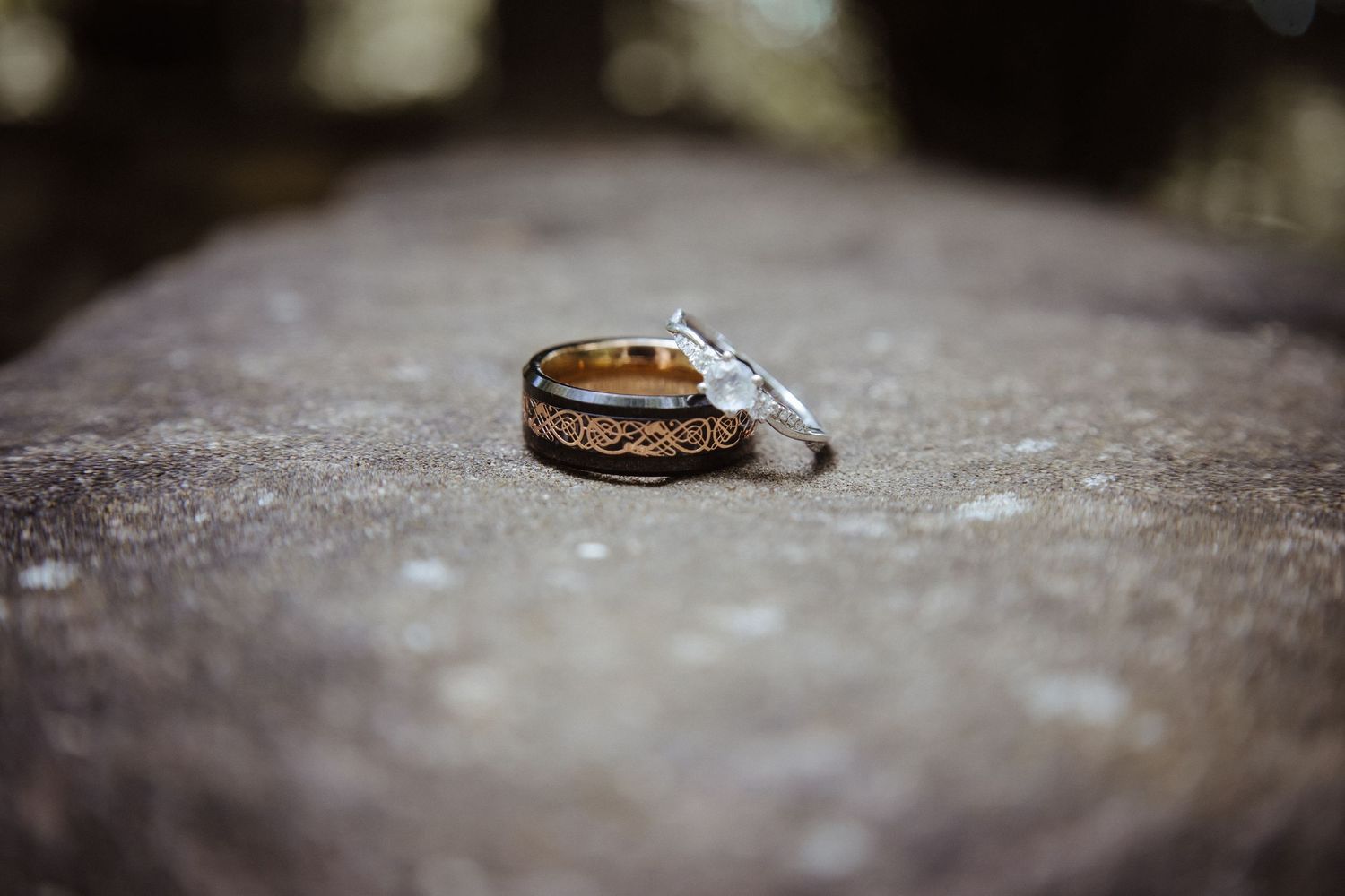 Two wedding rings, a gold band with intricate Celtic design and a diamond ring, resting on a stone surface.