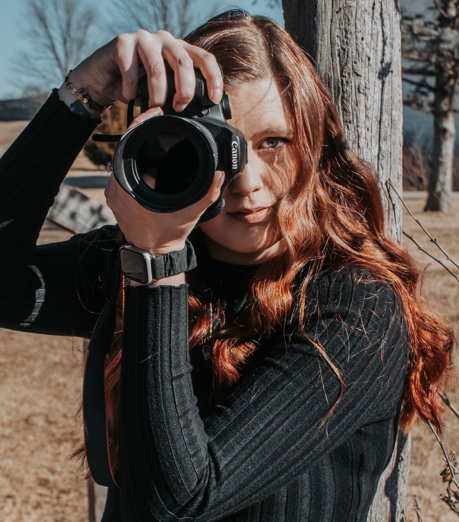 Young woman with red hair holding a Canon camera up to her eye outdoors near a tree.