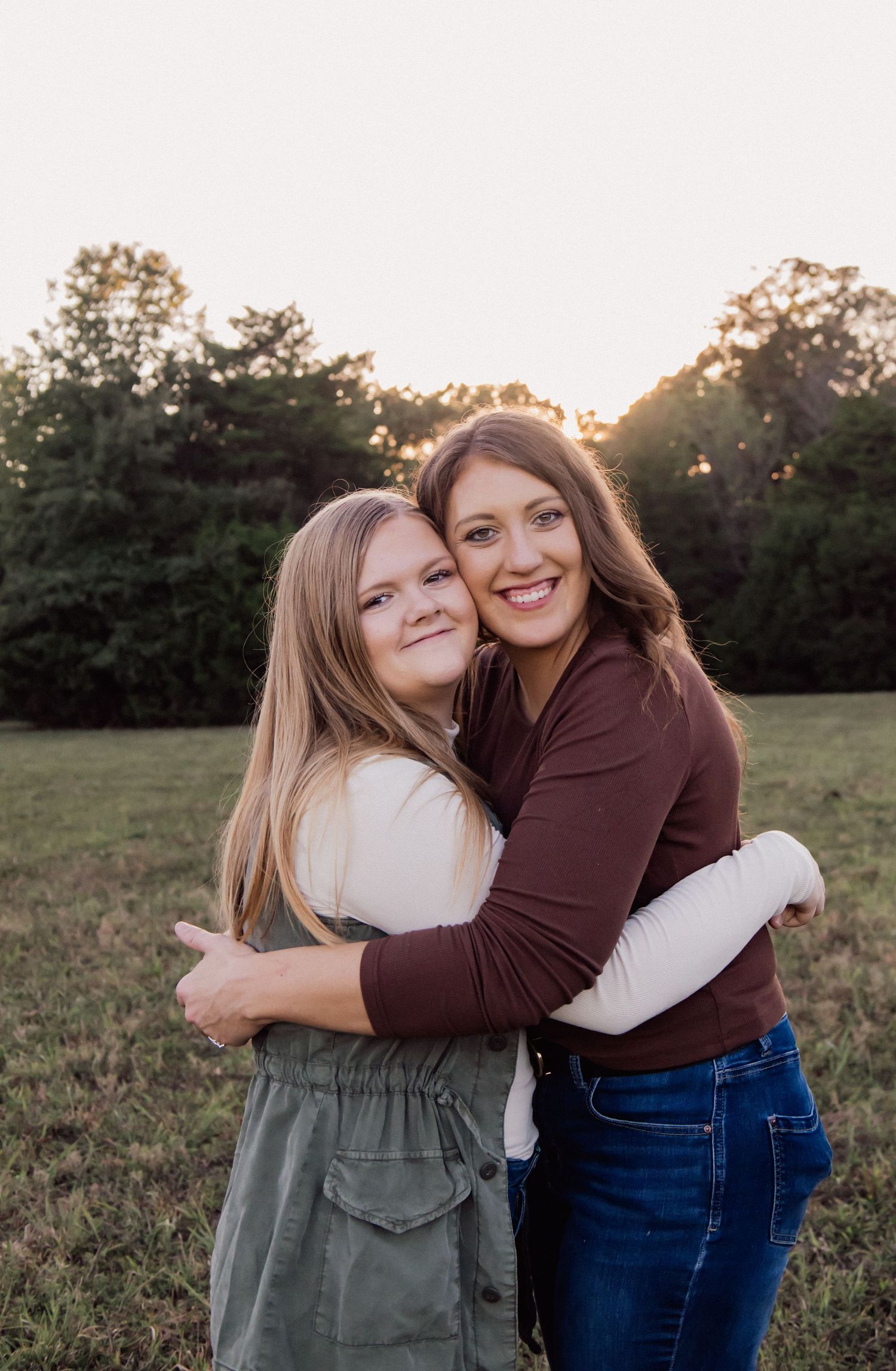 Two people embracing and smiling together in a field at sunset with trees in the background.