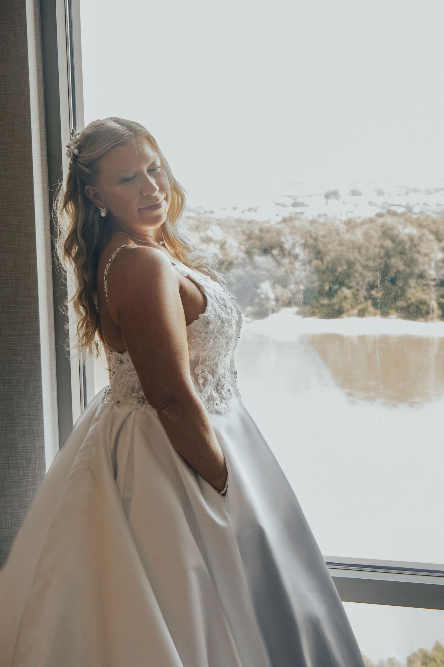 A bride in a white wedding gown laughs joyfully while standing near a window with natural light streaming in.