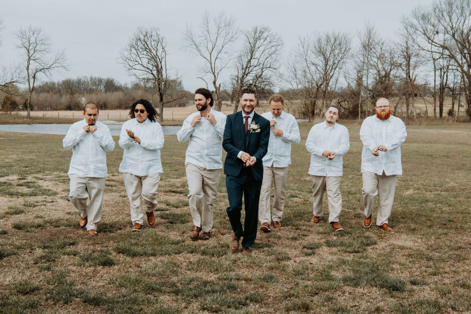 Wedding party walks together through a grassy field on a cloudy day wearing coordinated outfits in navy and beige.