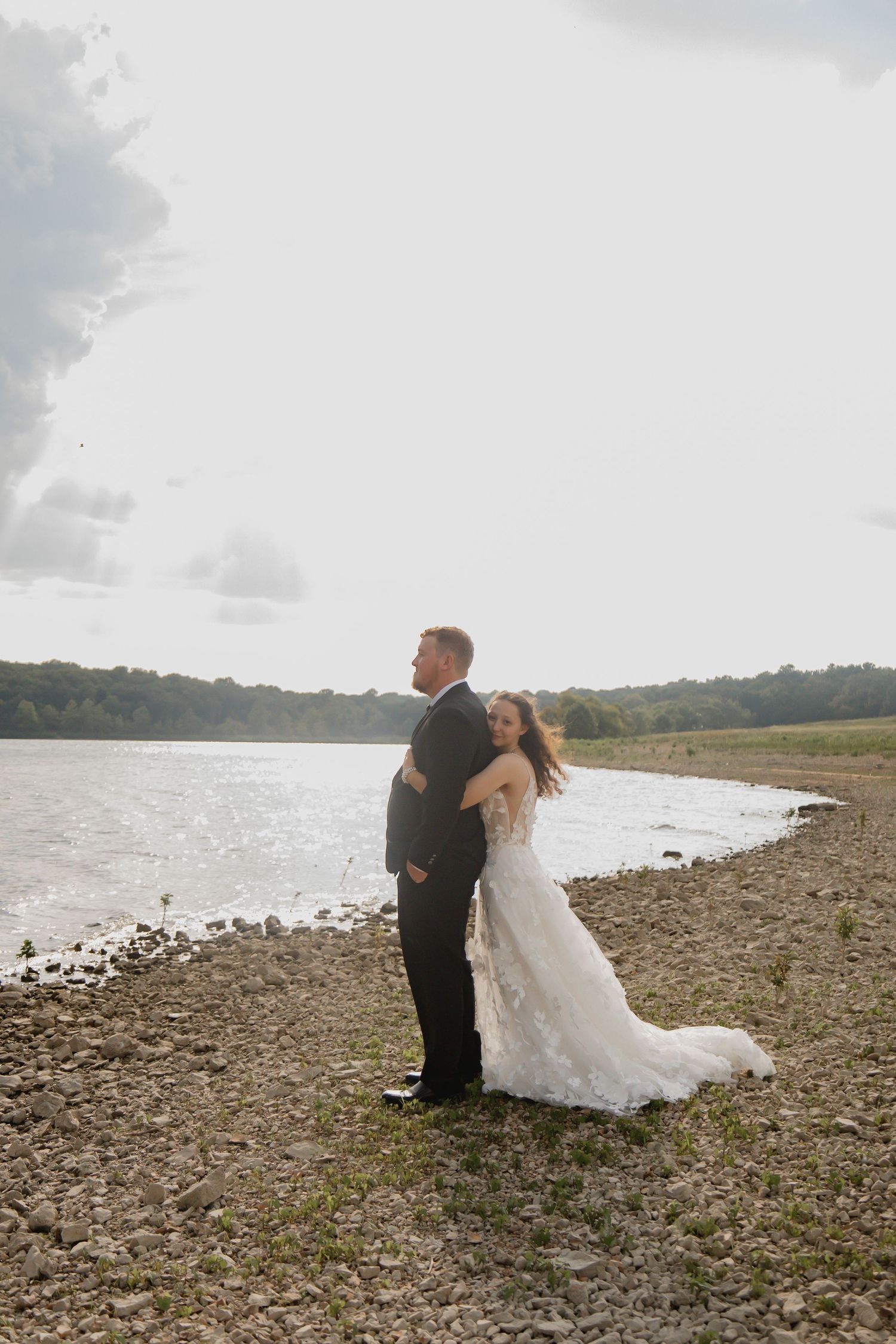 A couple shares a romantic moment on a rocky beach shoreline with water and trees in the background.