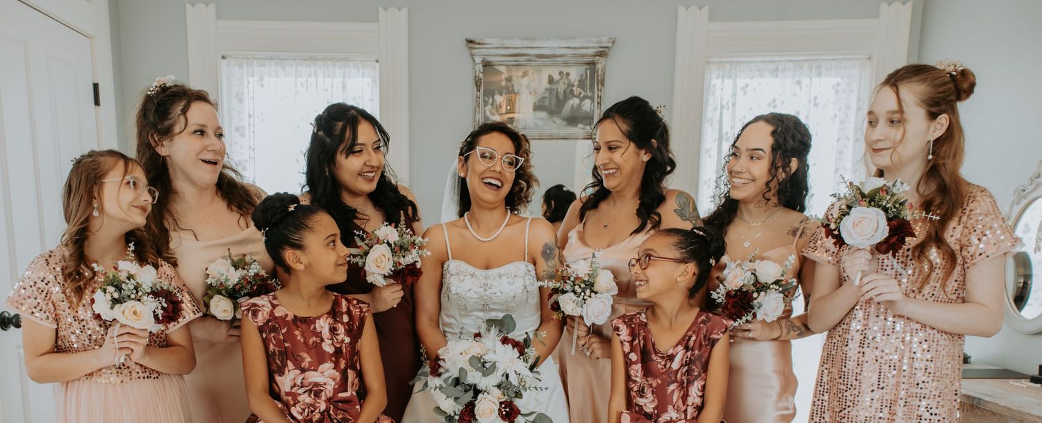 A group of bridesmaids in floral and pink dresses share a candid laughing moment with the bride at a wedding.