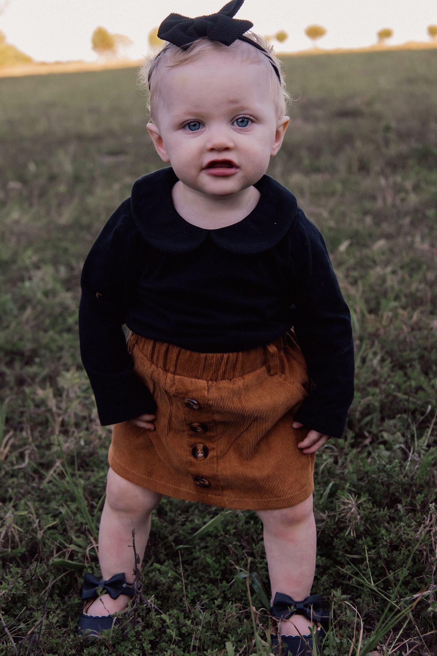A small child wearing a black and brown outfit plays on grassy ground while wearing blue sandals.