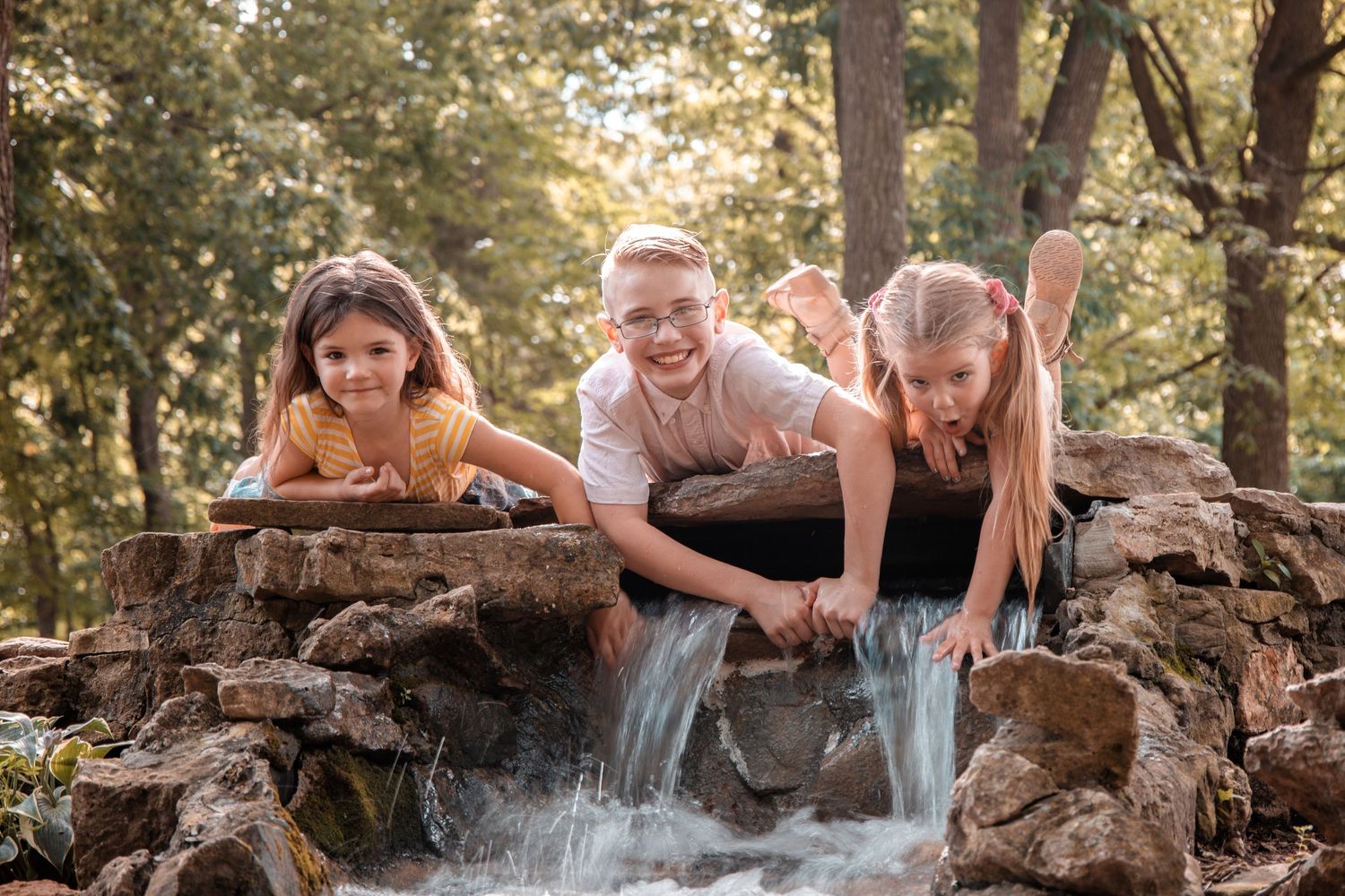 Three people laying on rocks near a small waterfall in a wooded setting during golden hour.