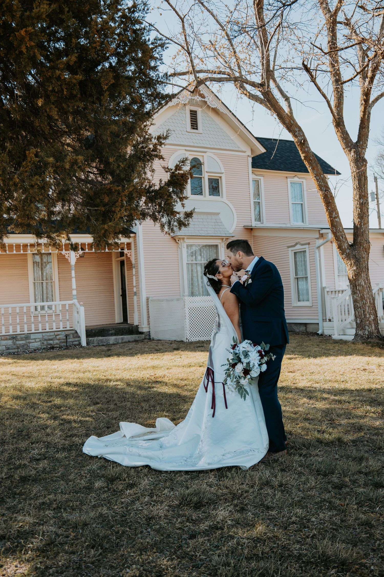 Romantic wedding portrait in front of a vintage pink Victorian house with trees framing the scene.