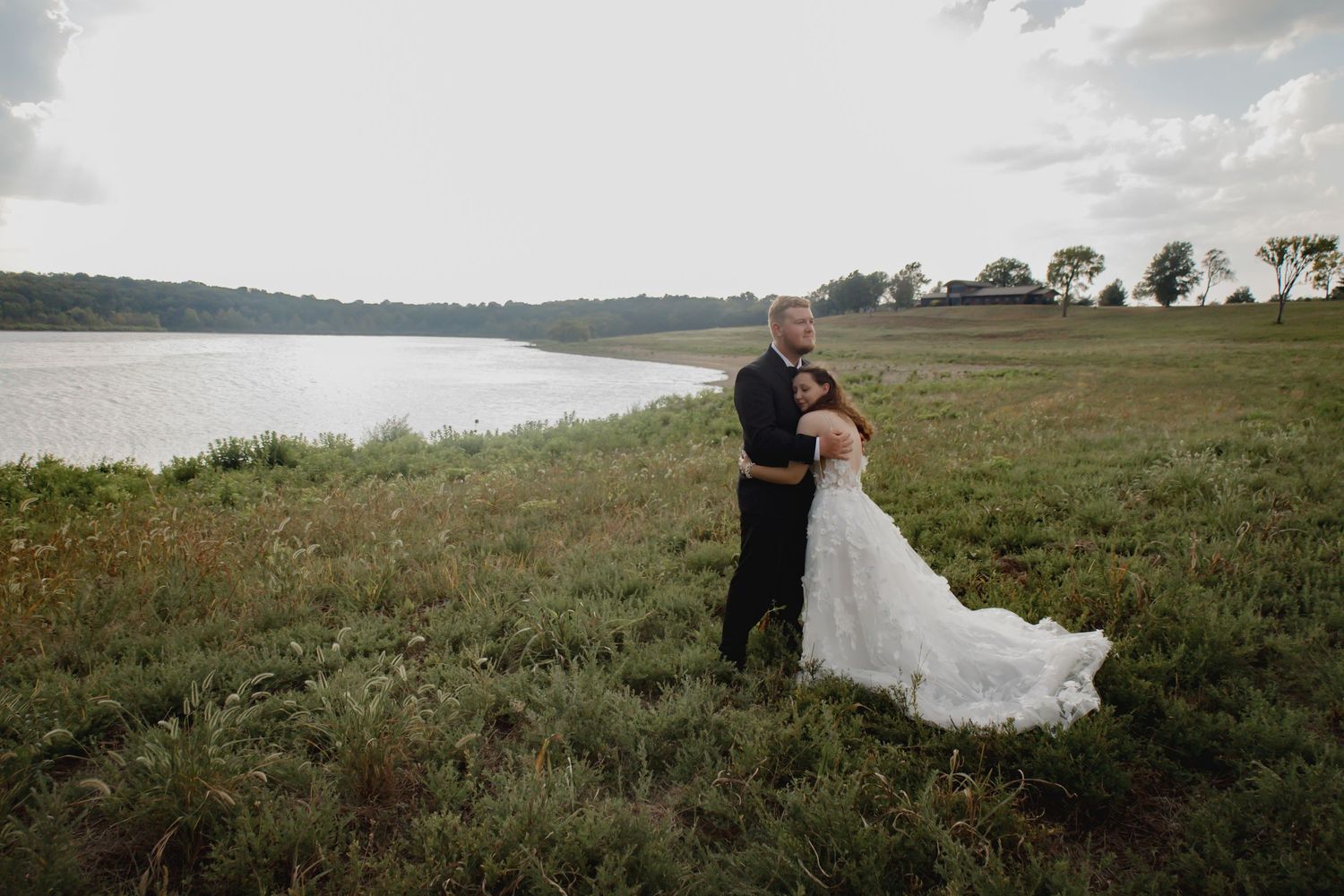 A couple in wedding attire stands together on grassy terrain overlooking a serene lake.