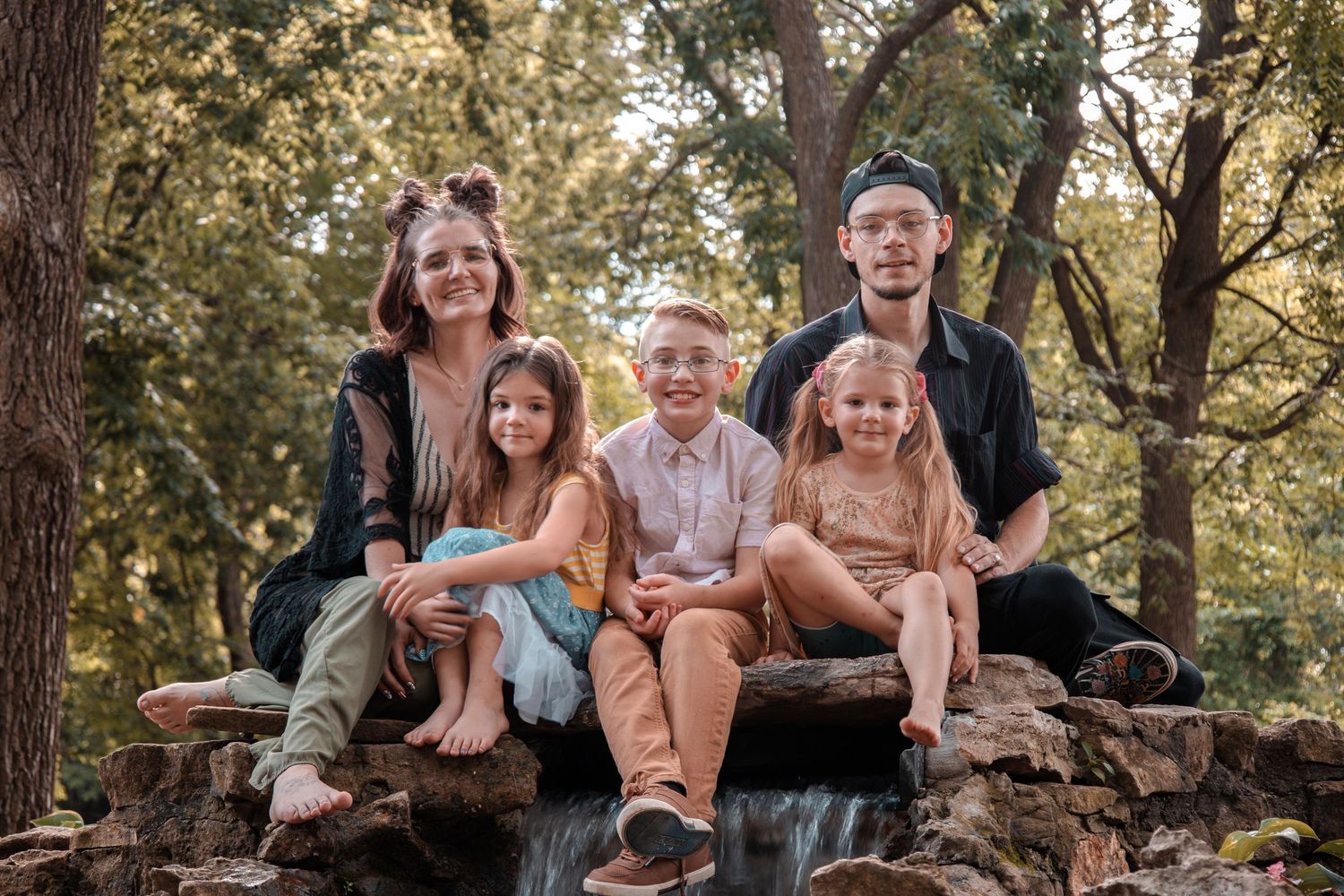 A family sits together on a rustic stone wall surrounded by autumn trees during an outdoor photo session.