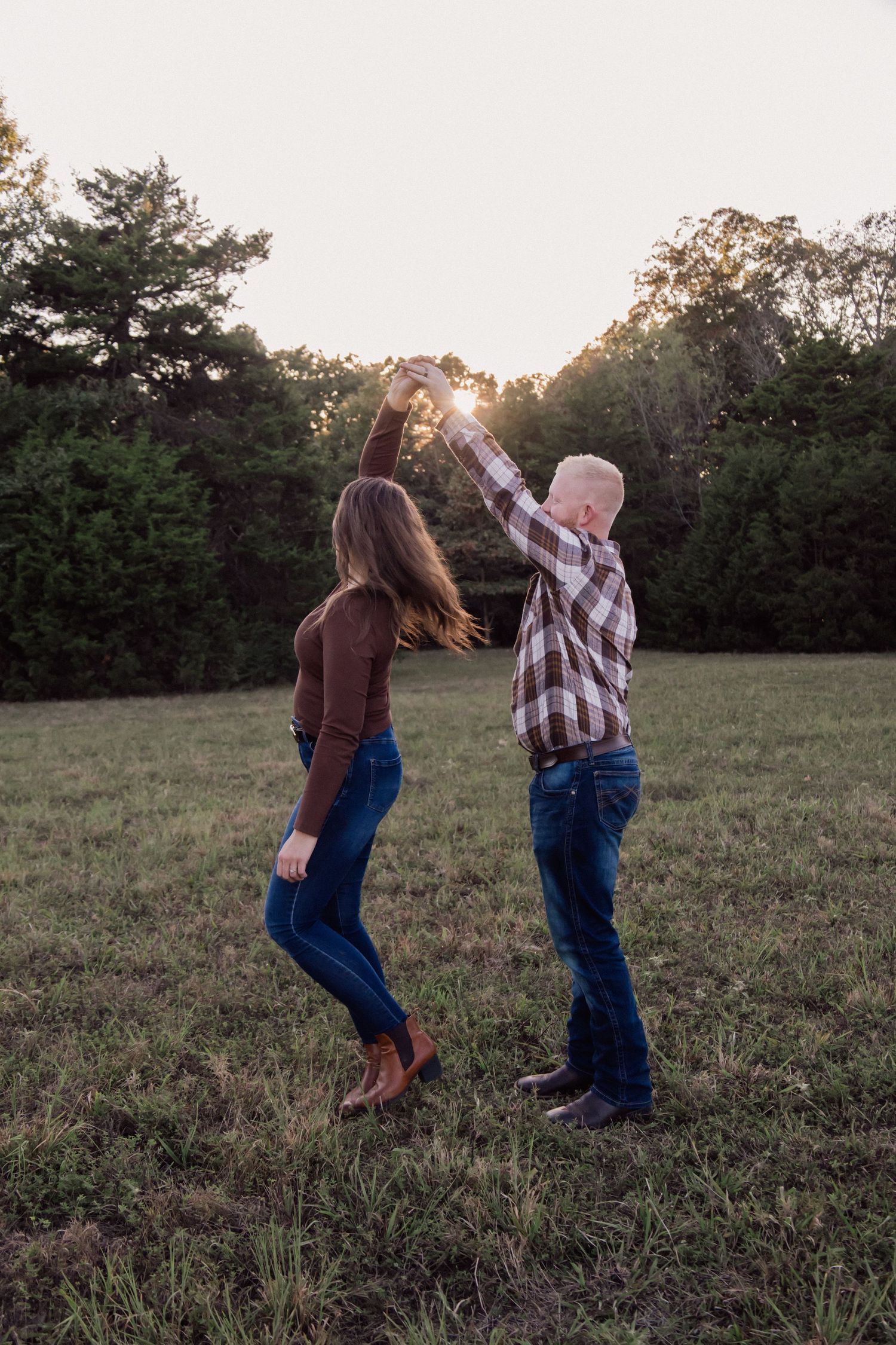 Couple dancing in a sunlit field at golden hour, wearing jeans and plaid, surrounded by lush green trees.