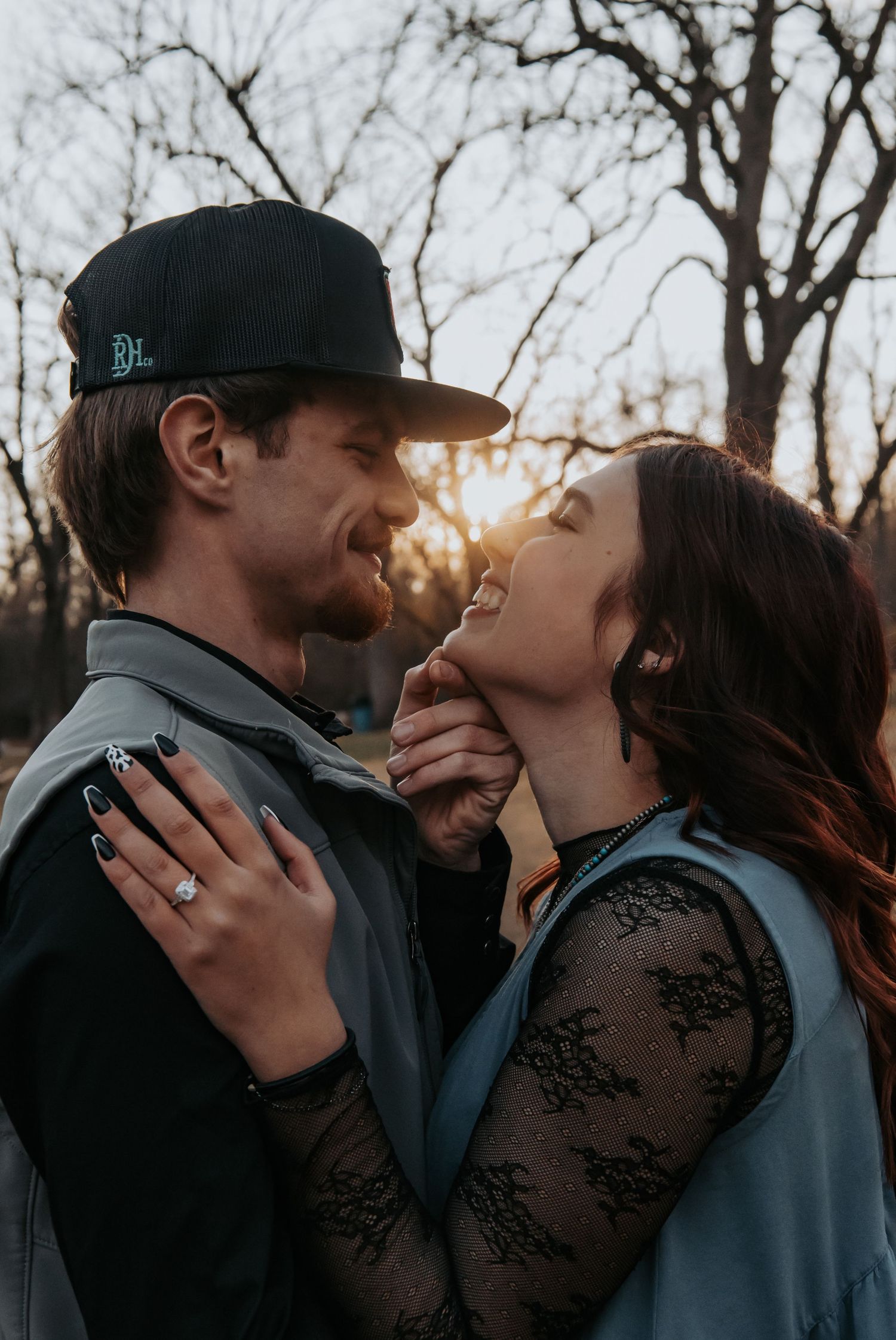 Couple sharing a romantic moment at sunset, woman showing engagement ring, bare trees in background.
