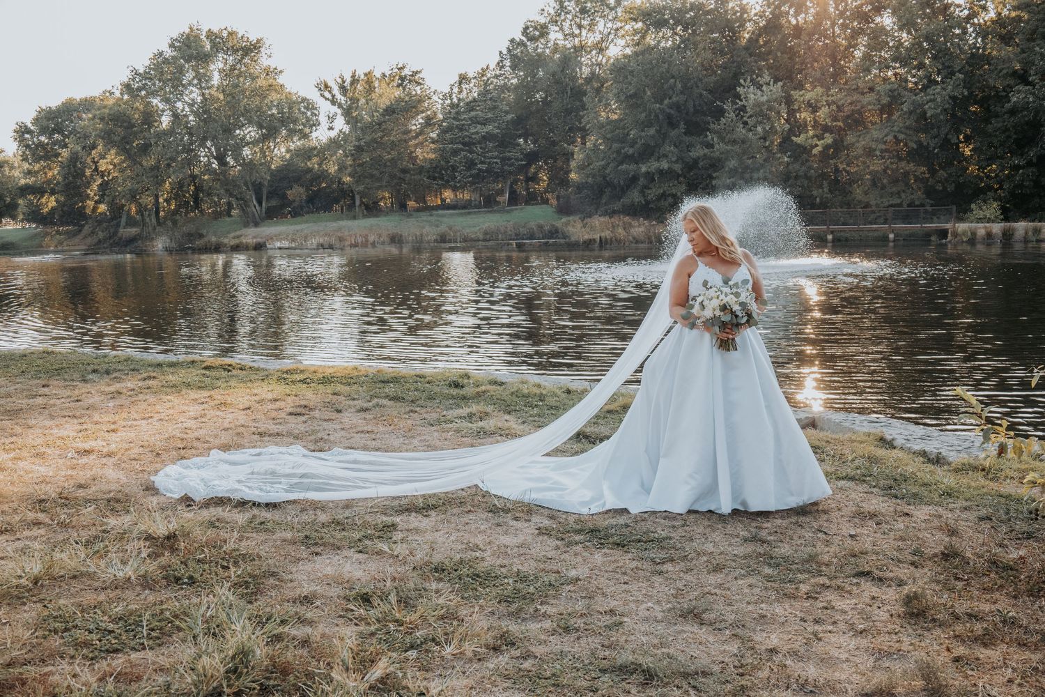 A flowing white wedding dress with a long cathedral veil creates an ethereal silhouette against a lakeside backdrop at sunset.