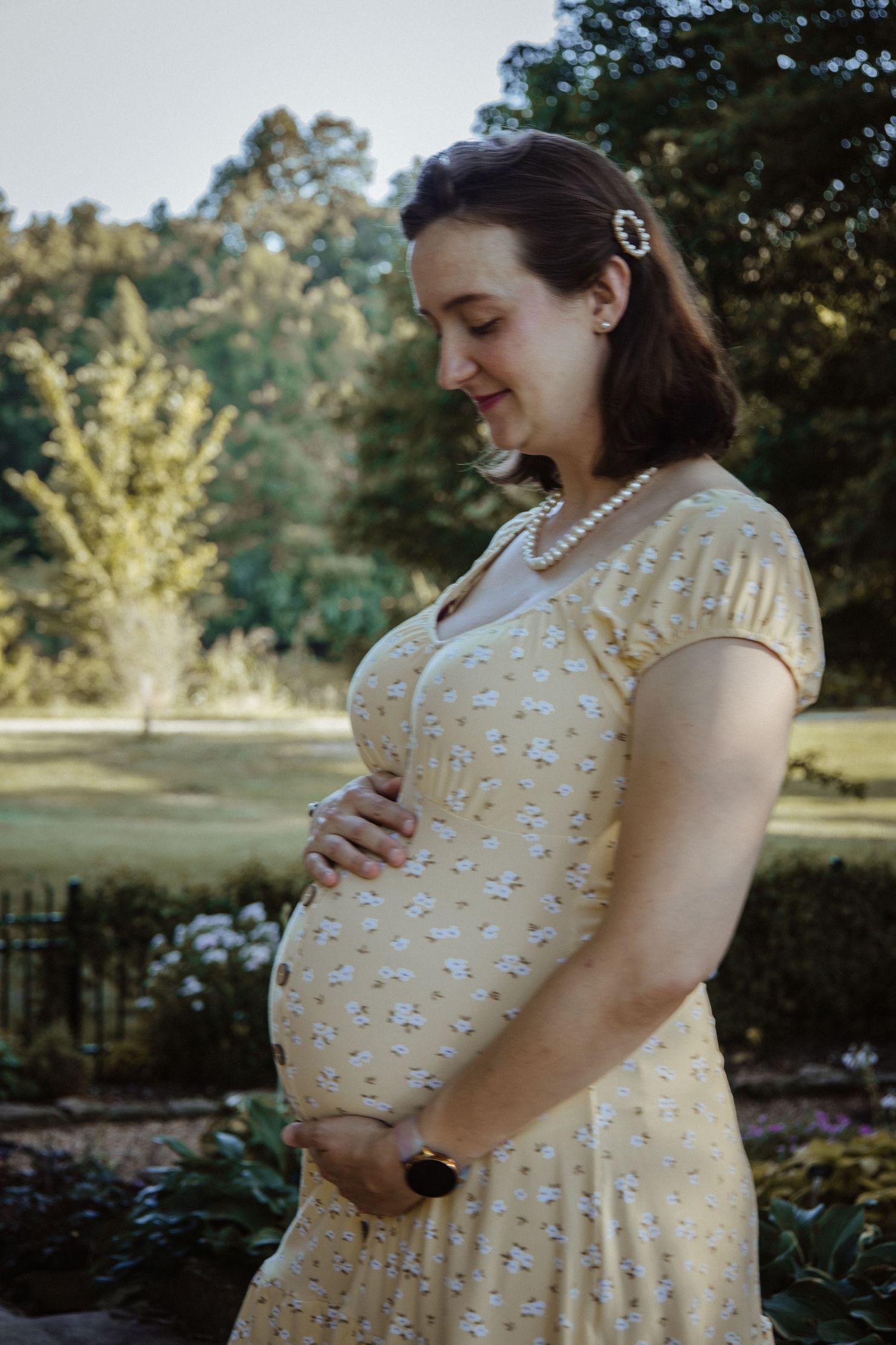 A pregnant figure in a yellow floral dress with pearl necklace stands in a garden setting during golden hour.