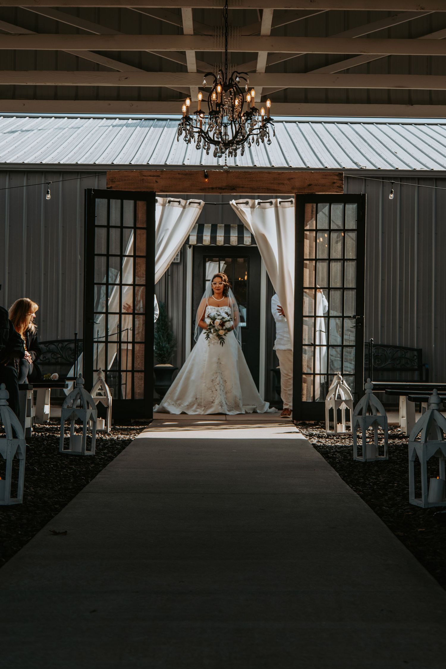 An elegant wedding ceremony setup with a black runner, lanterns, and chandelier leading to a draped doorway entrance.