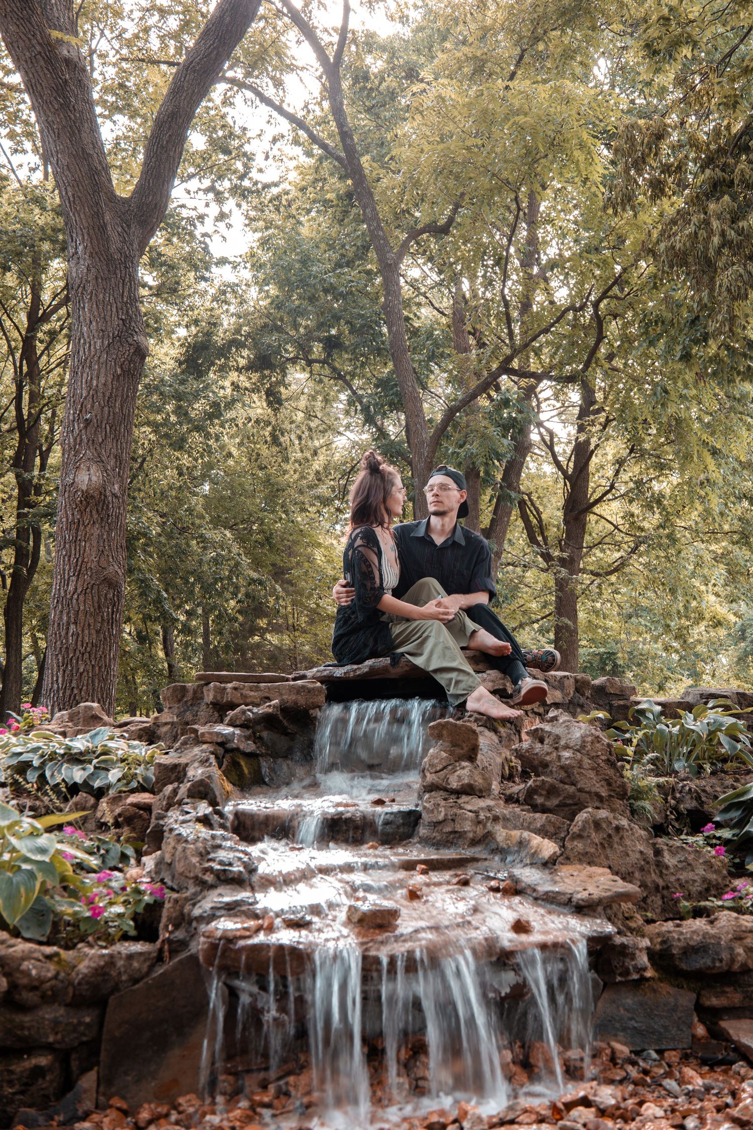 A couple sits together on rocky ledge by cascading waterfall in a serene forest setting.