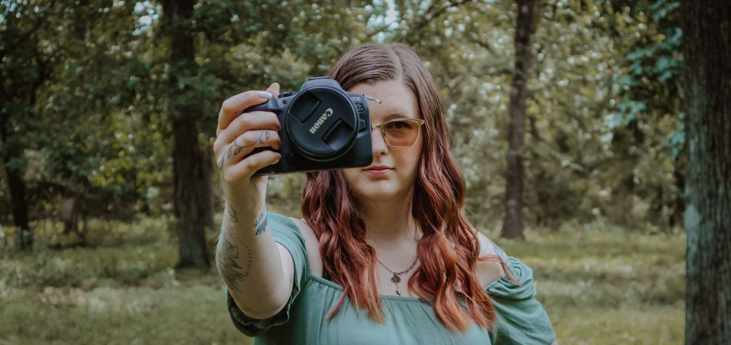 A person with red hair takes a selfie with a DSLR camera while standing outdoors in a natural setting.