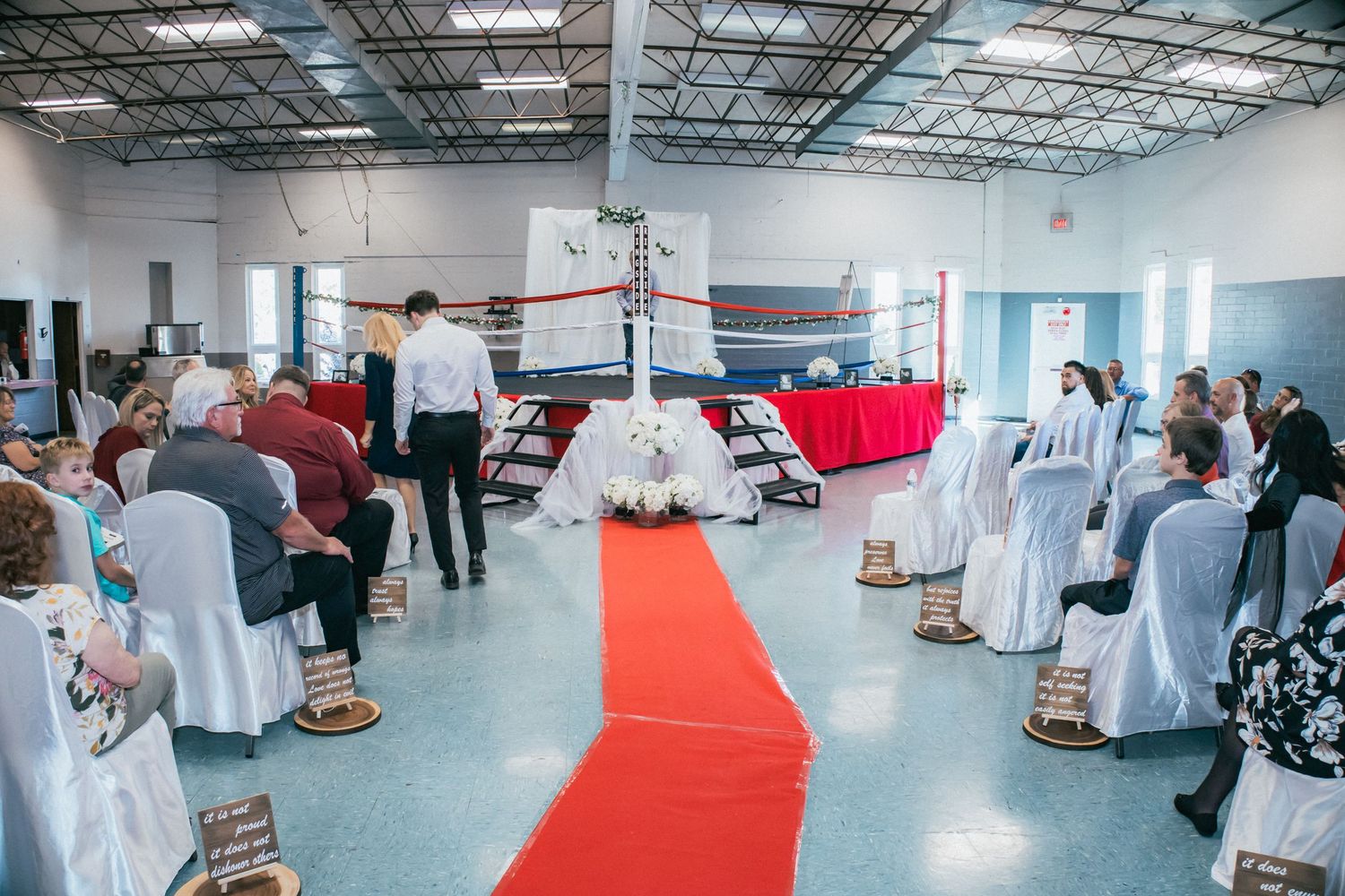 People gather in a spacious event venue with white chairs and red carpet for a business or networking function.