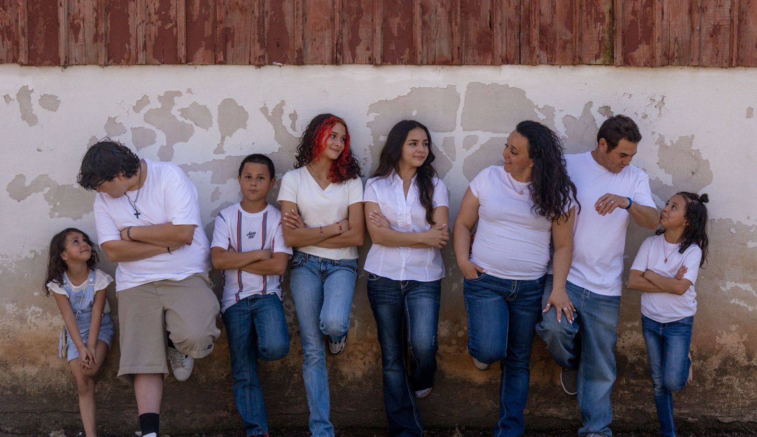 Family of eight in white shirts and jeans posing casually against a weathered wall outdoors.