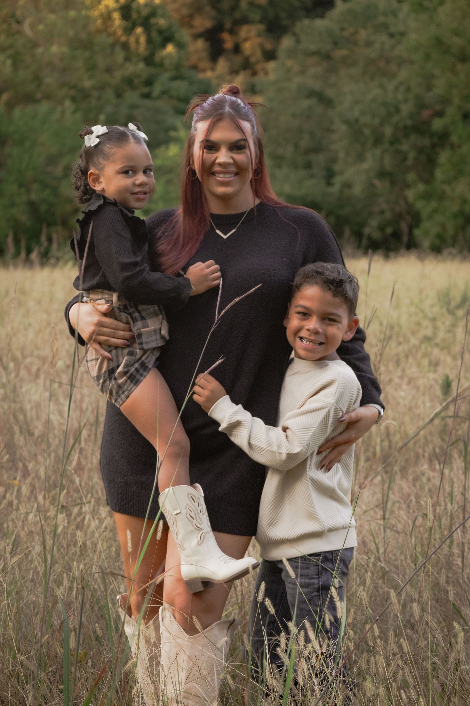 A family poses together in a tall grass field during golden hour wearing coordinated black and cream outfits.