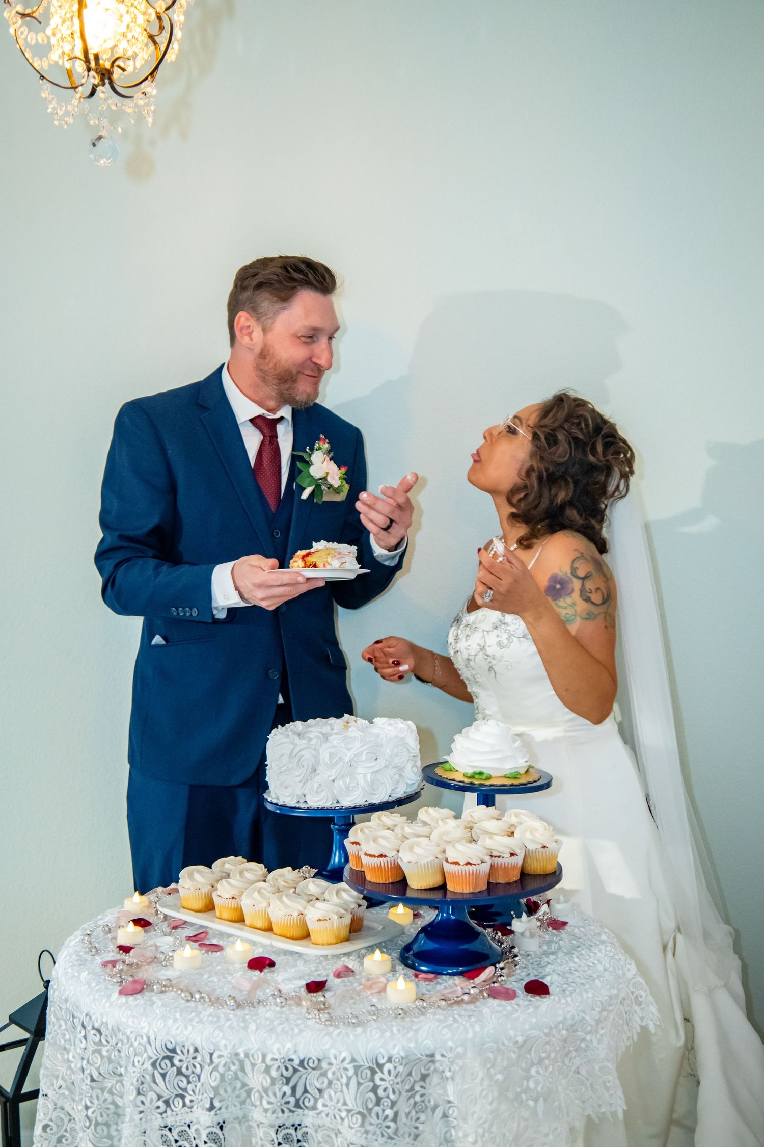 A wedding couple sharing their first bite of cake at an elegant dessert table with cupcakes and white lace tablecloth.