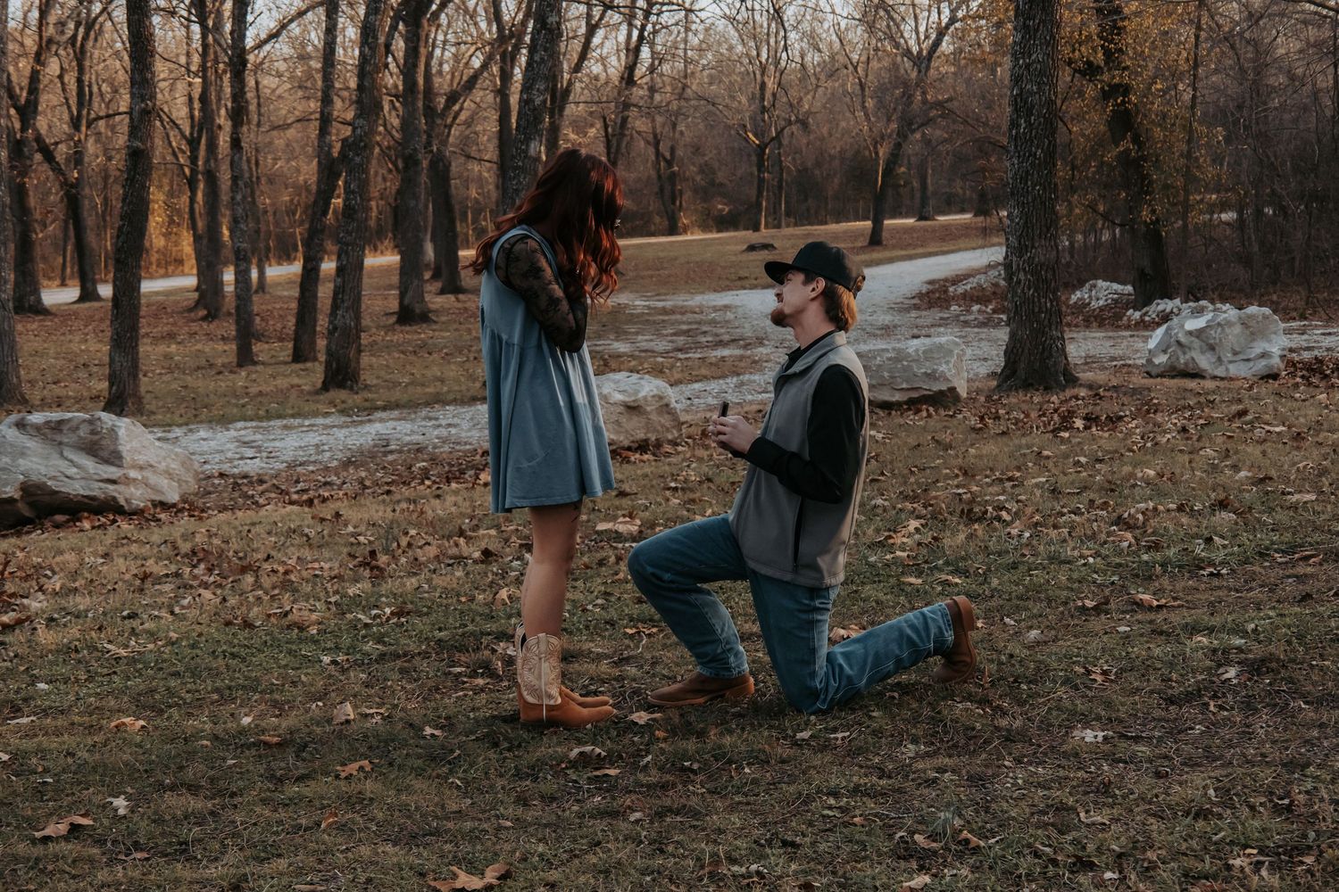 A couple in casual attire sits together on fallen autumn leaves in a wooded park setting during golden hour.