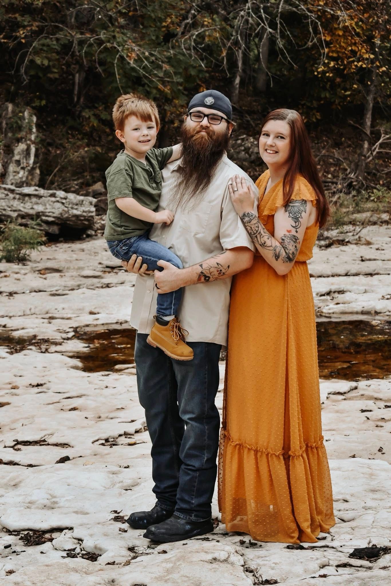 Family portrait on rocky terrain with woman in mustard maxi dress, bearded man in casual wear, and young child in green shirt.