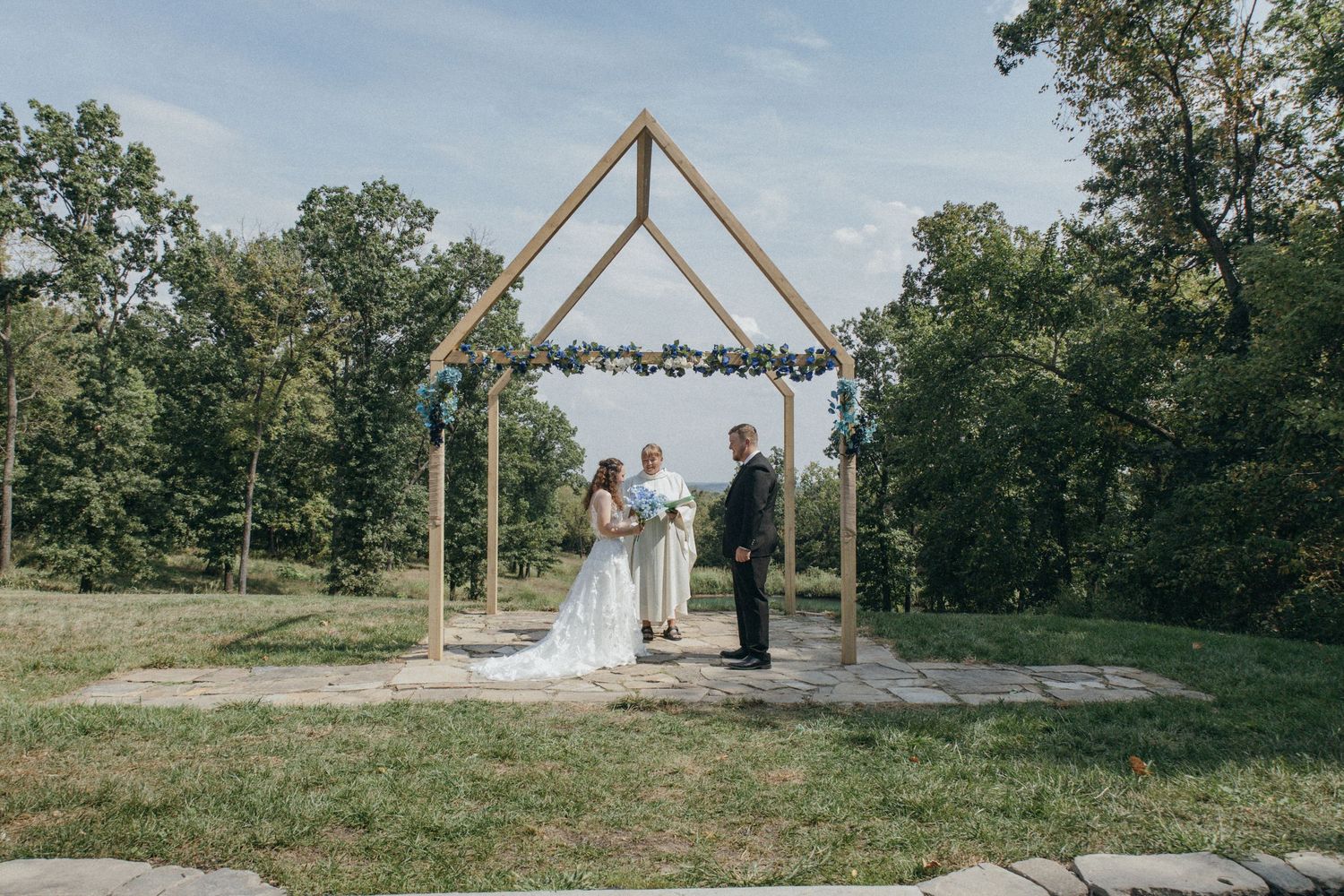 A rustic outdoor wedding ceremony setup with wooden triangle arch and chairs on a hilltop overlooking nature.
