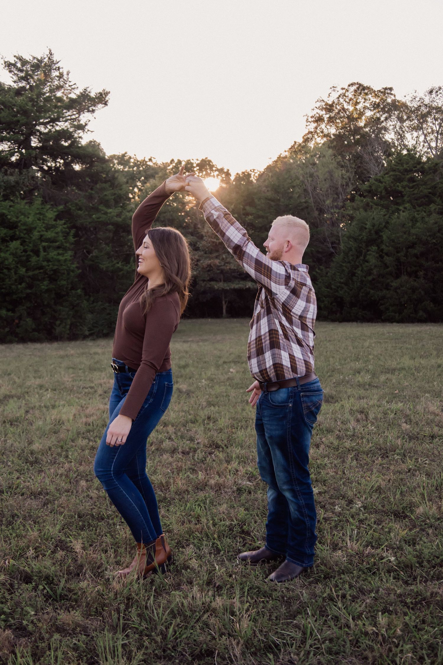 A couple dances together in a field at sunset with trees in the background.