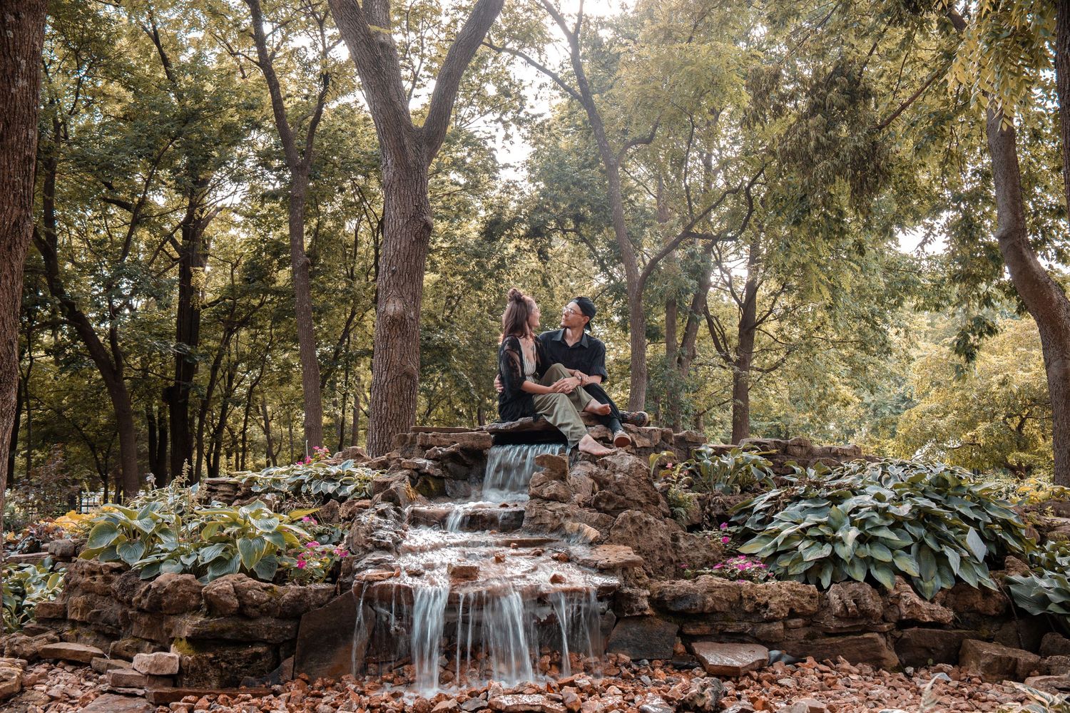 A couple sits together on a scenic stone waterfall surrounded by lush greenery in a wooded park setting.