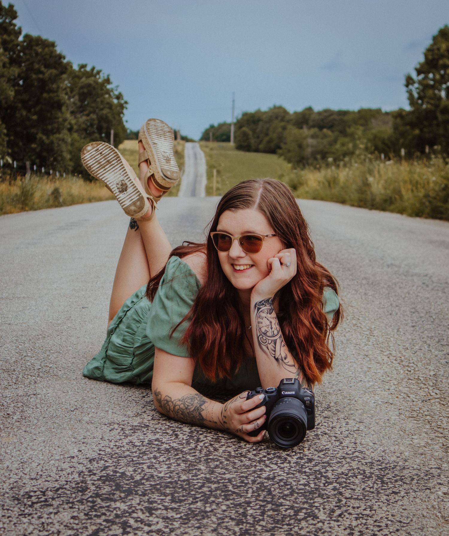 Photographer lying on empty rural road with camera and boots, wearing green shirt and sunglasses on sunny day.