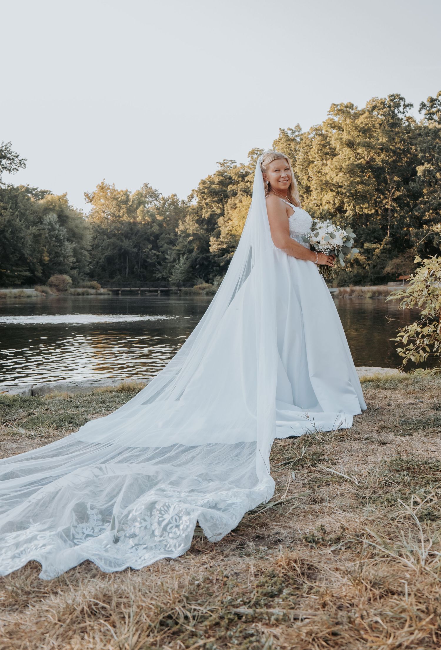 A stunning bridal portrait showcasing an elegant white wedding gown with a dramatic cathedral-length veil by a serene lake at sunset.