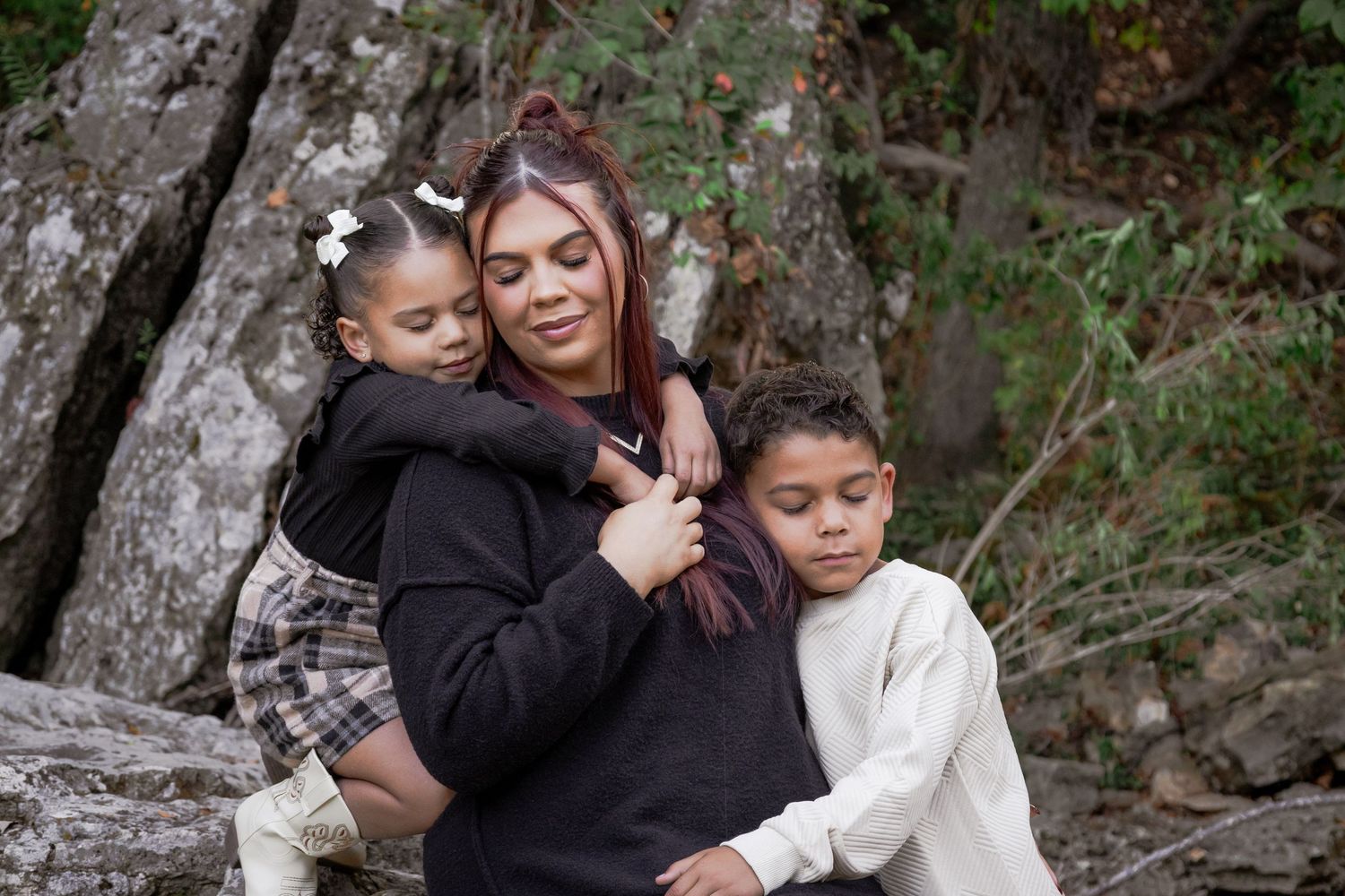 A family shares warm embraces while posing outdoors against a natural backdrop of trees and rocks.