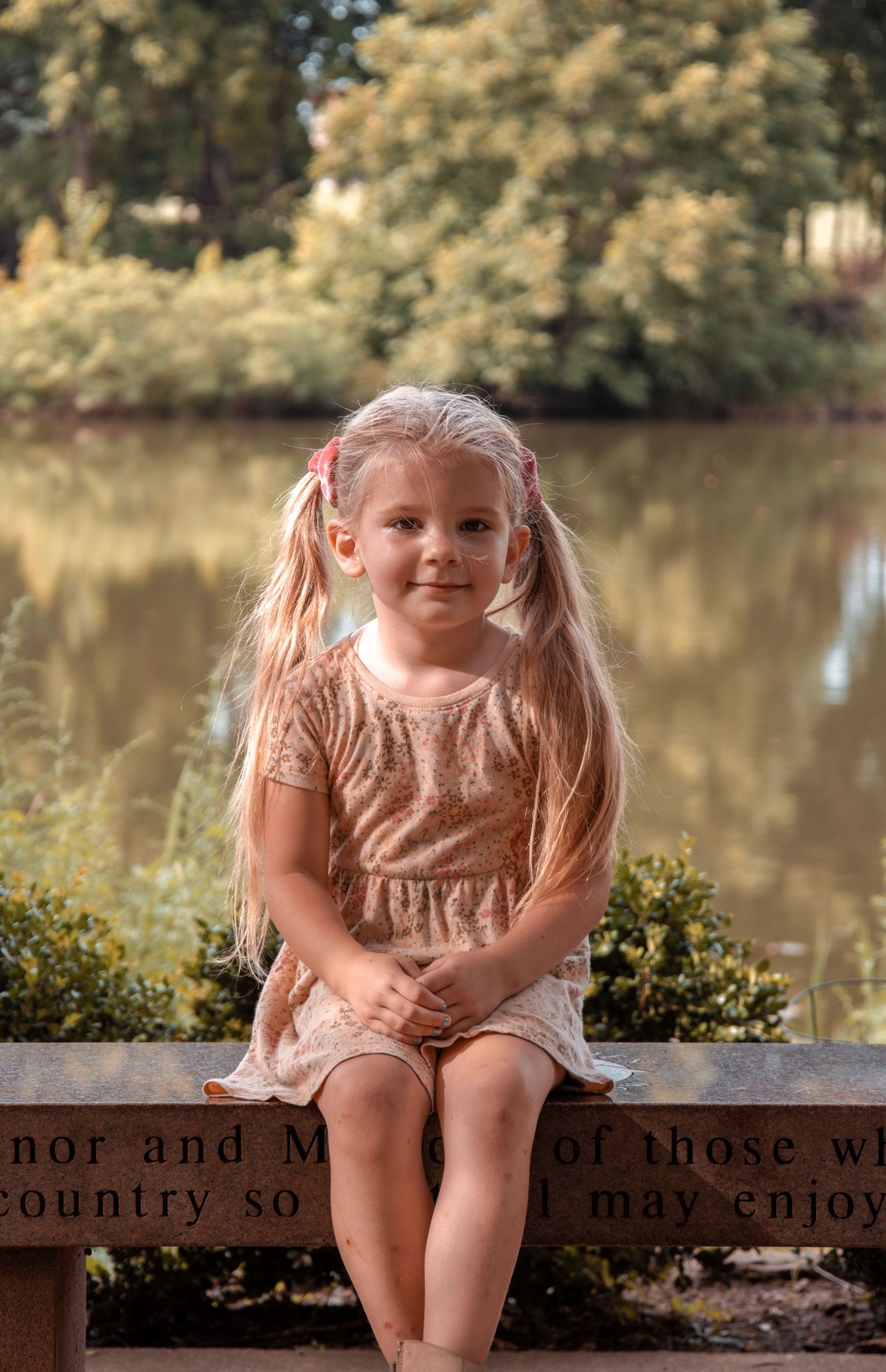 A child in a beige dress sits on a wooden bench near a tranquil pond with trees in the background.