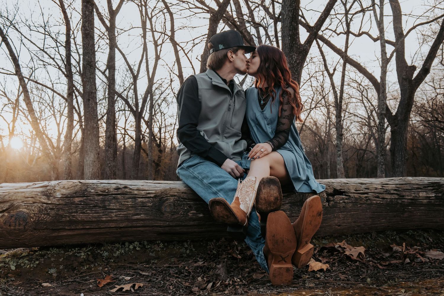 A couple sits together on a fallen tree trunk in a moody winter forest setting during golden hour.