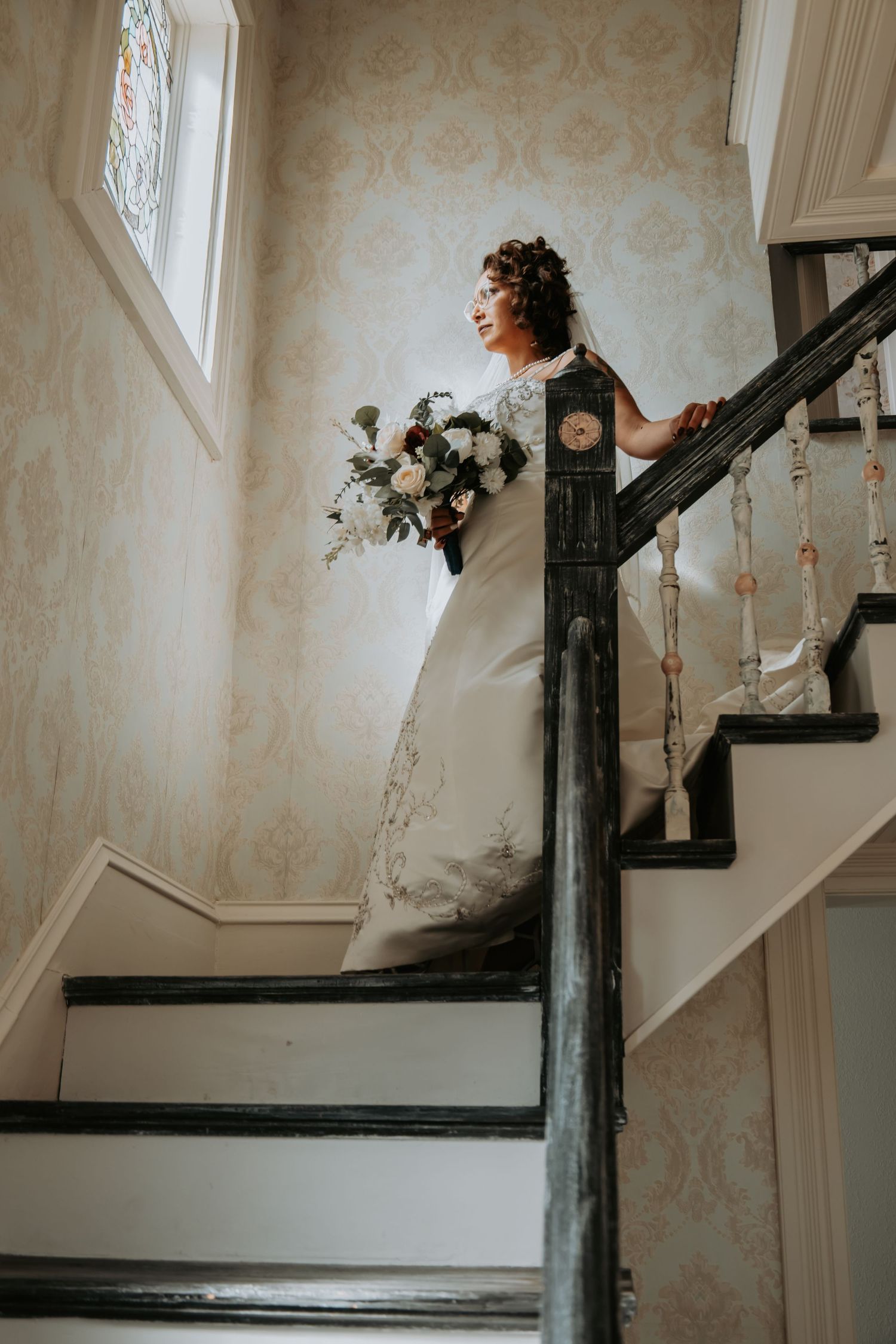 A bride in a flowing white dress stands on vintage stairs holding a lush bouquet against floral wallpaper in soft lighting.