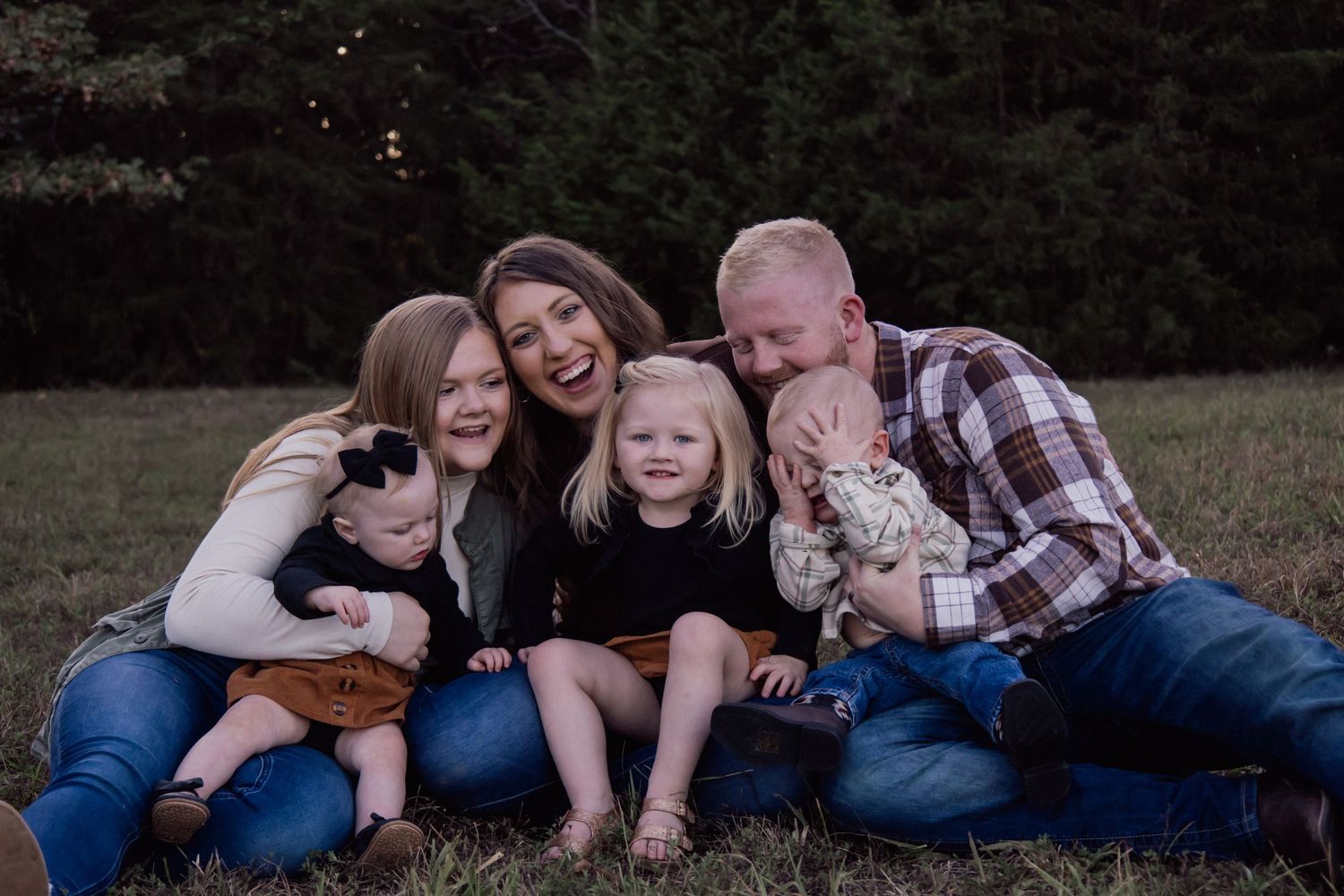 A family sits together on a grassy hillside during golden hour, creating a warm and intimate outdoor portrait.