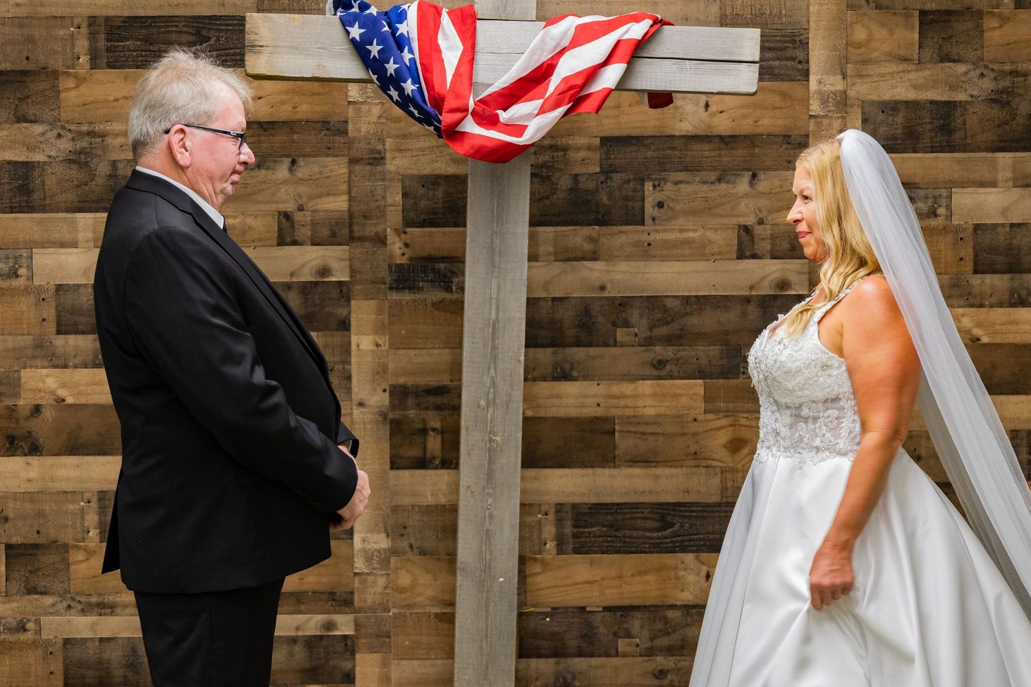 A wedding couple and officiant stand before a rustic wooden wall backdrop with American flags during their ceremony.