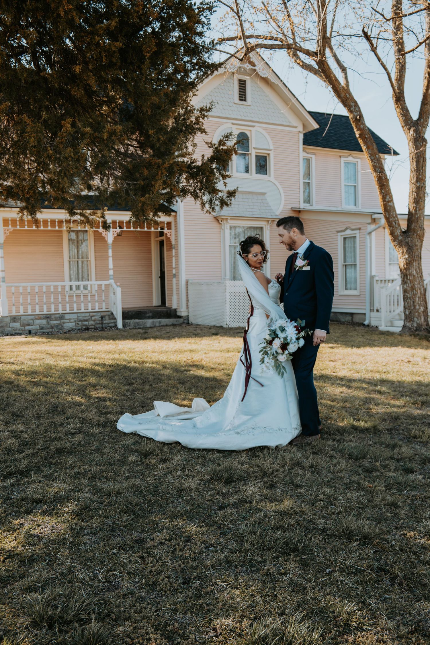 Wedding couple stands in front of a vintage Victorian-style house with white trim and wraparound porch on a winter day.