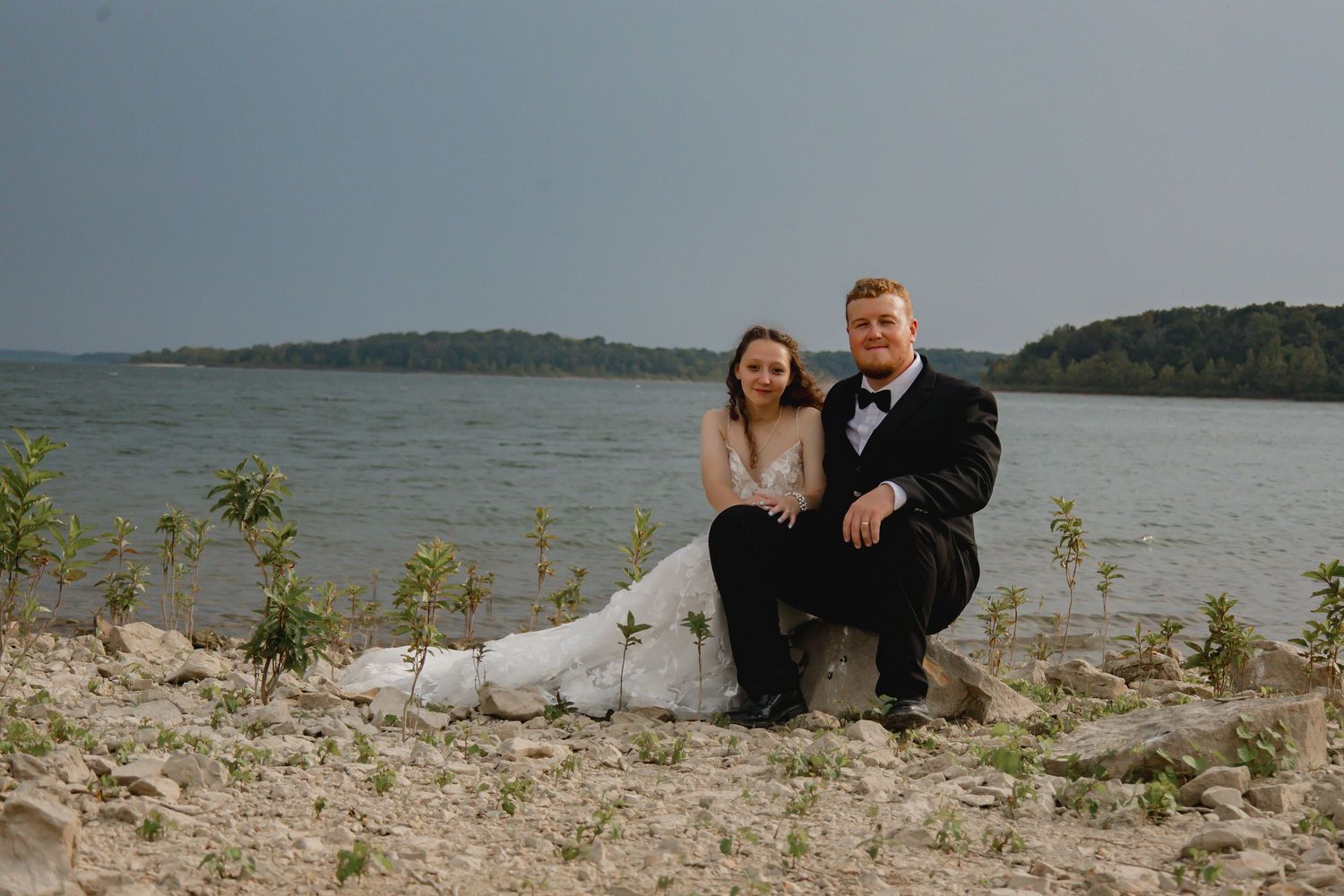 A couple sits together on a rocky shoreline overlooking a scenic lake on a cloudy day.